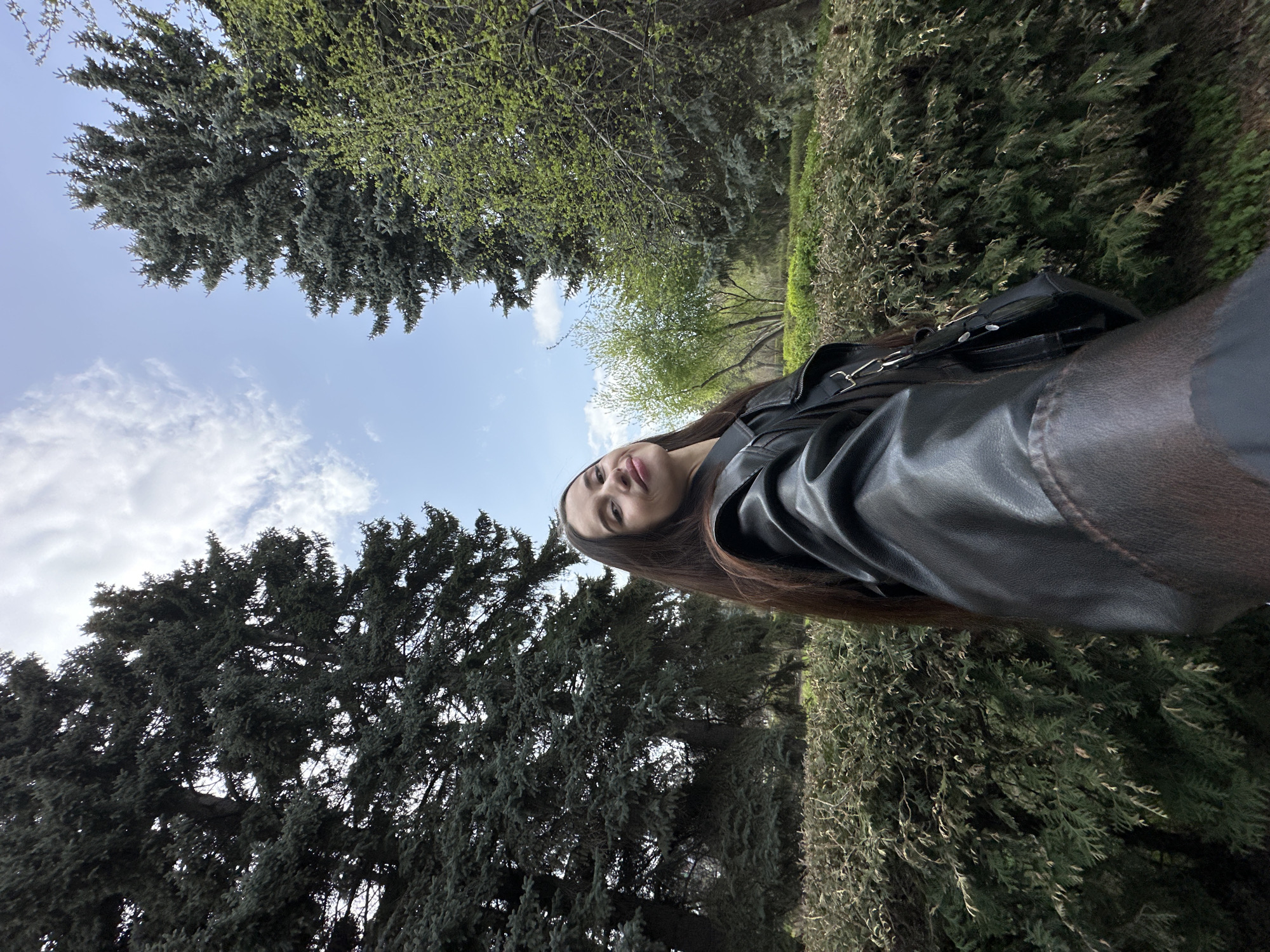 A low-angle portrait captures a young woman standing outdoors in what appears to be a park or garden in Yekaterinburg, Russia. She is positioned on the right side of the frame, looking upwards and directly at the viewer with a neutral expression. She has long, dark hair and is wearing a black leather-like jacket.
The background is filled with natural elements. To the left and upper portion of the image are tall, dense evergreen trees with dark green needles. Other trees with lighter green, possibly new spring foliage, are also visible. A dense row of green bushes or a trimmed hedge extends horizontally across the mid-ground. Above the trees, the sky is a bright blue with scattered, wispy white clouds, indicating clear to partly cloudy daytime weather. The overall impression is one of natural beauty and an early to mid-day setting. No specific activity is depicted other than posing for the photograph. There is no visible text.