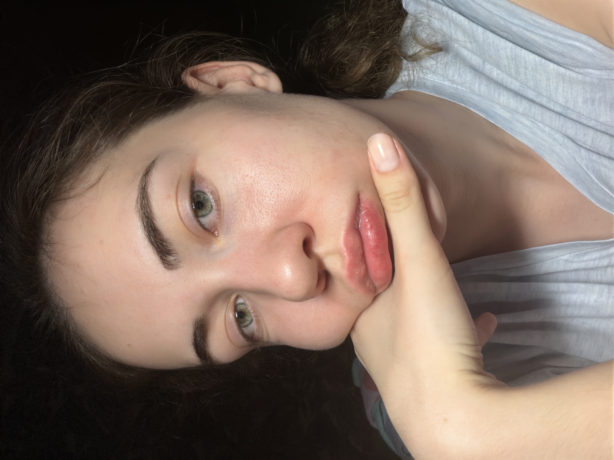 A close-up, waist-up, head-and-shoulders portrait of a young woman with light brown hair and green eyes is depicted against a dark, out-of-focus background. She is resting her chin and jawline on her right hand, with her fingers extended. The woman is wearing a light blue t-shirt. Her left cheek appears unusually swollen. Her lips are slightly parted, and her expression is neutral. The lighting is frontal and appears to be artificial. There is no discernible indication of the location being Zavodoukovsk, Russia.