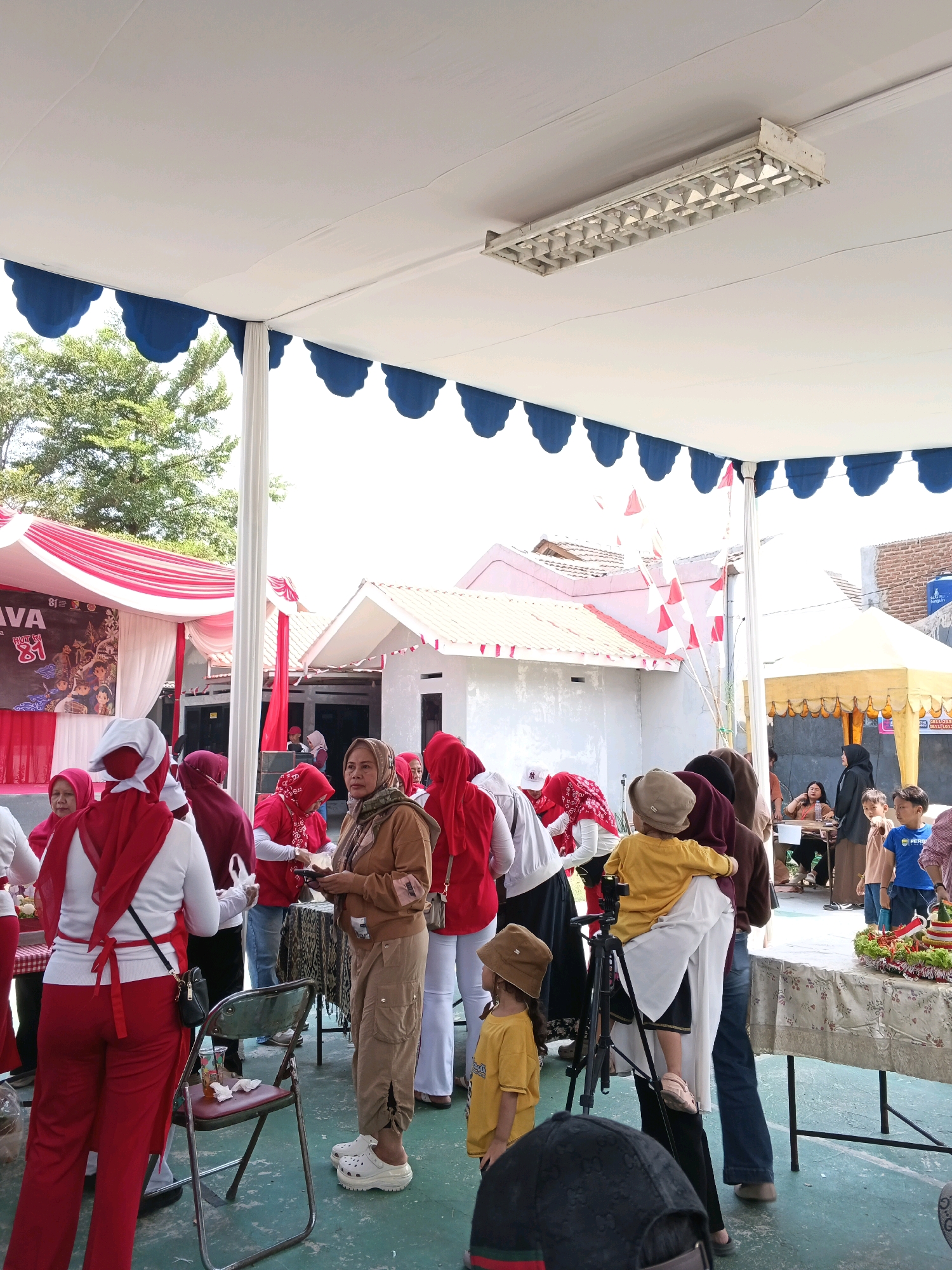 The image depicts an outdoor event, possibly a festival or market, under a white tent with blue scalloped trim. The scene is set in Margahayu, Indonesia. A group of people, mostly women wearing red and white attire, are gathered. Some are interacting, appearing to be serving or receiving items from a stall. In the foreground, a woman in a brown hooded jacket and beige cargo pants stands near a folding chair. To her right, a child in a yellow shirt stands, facing away from the camera. In the background, there are several buildings with red roofs and white walls, and a yellow tent. Flags, likely red and white Indonesian flags, are visible. The ground is a green paved surface. The overall atmosphere suggests a community gathering or celebration.