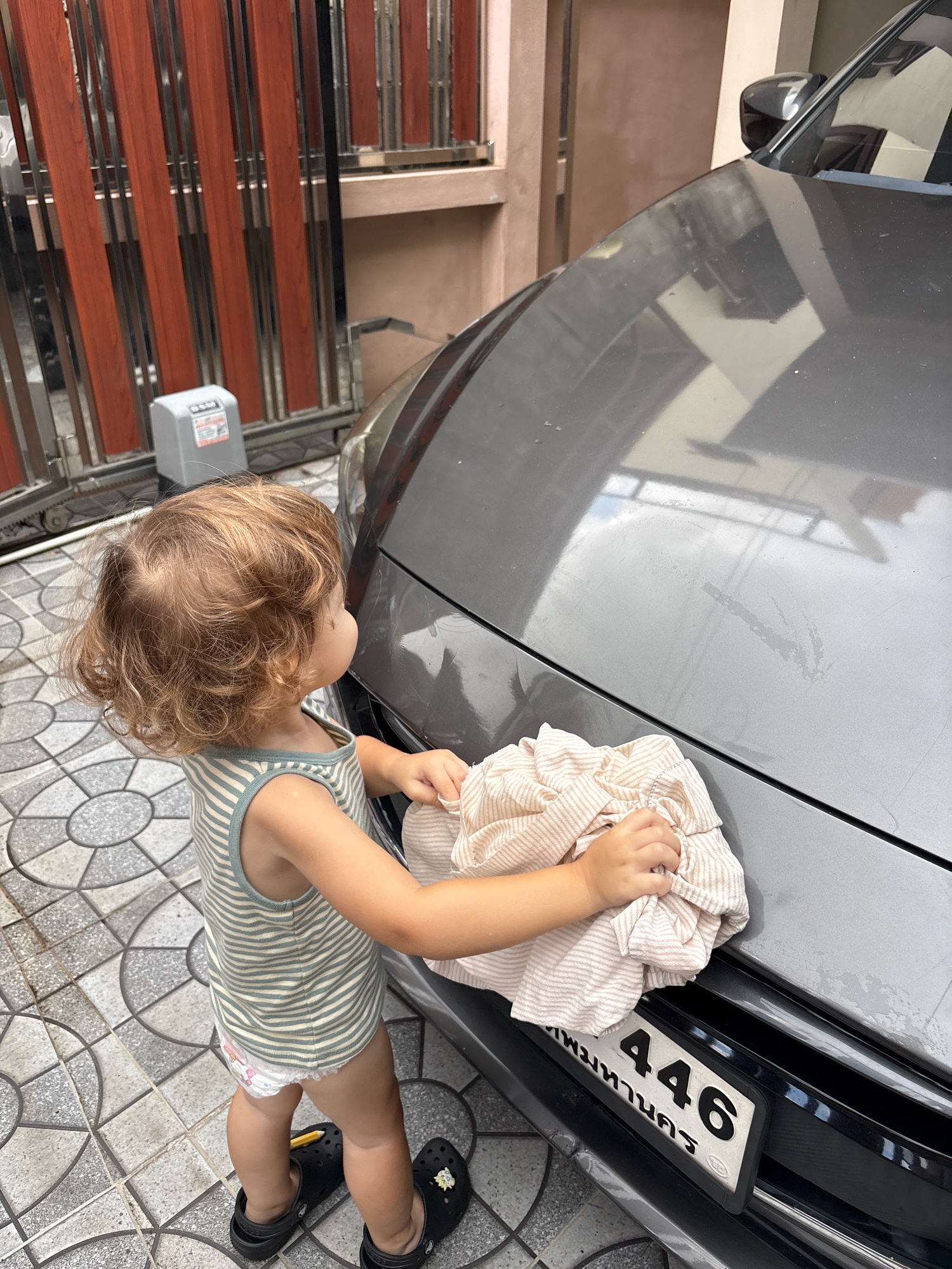 A toddler with curly blonde hair, wearing a striped tank top and a diaper, is standing on a tiled driveway in Choeng Thale, Thailand, and wiping a dark gray car with a light-colored striped cloth. The child is facing away from the camera, looking towards the car's hood and fender. A license plate with the numbers "446" and Thai script is visible on the front of the car. In the background, a wooden and metal gate is visible, along with a gray gate motor. The scene appears to be outdoors, possibly in a residential area or a car wash.
