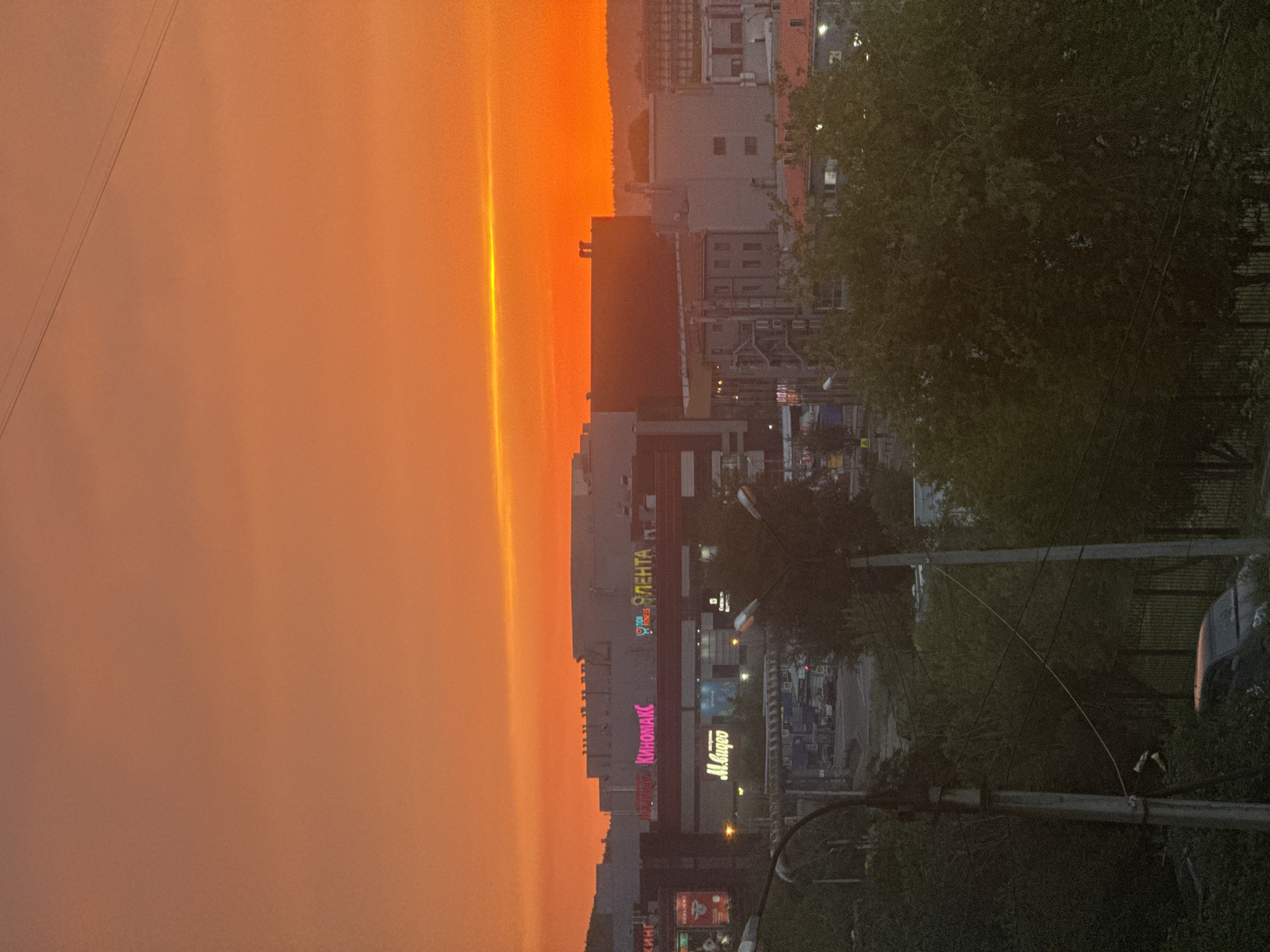 The image captures a city skyline at sunset in Irkutsk, Russia, dominated by a vibrant orange and yellow sky. In the mid-ground, a large commercial building complex is visible, featuring illuminated signs such as "КиноМАКС" (KinomaKS), "М.Видео" (M.Video), and "ЛЕНТА" (LENTA). The complex appears to be a shopping mall or entertainment center. Several smaller buildings and industrial structures are also present in the background. In the foreground, dense green trees and foliage obscure parts of the view. A street lamp and power lines are also visible. A dark-colored car is partially visible in the lower left corner, parked near a fence. The overall scene depicts an urban environment transitioning into evening under a dramatic sky.