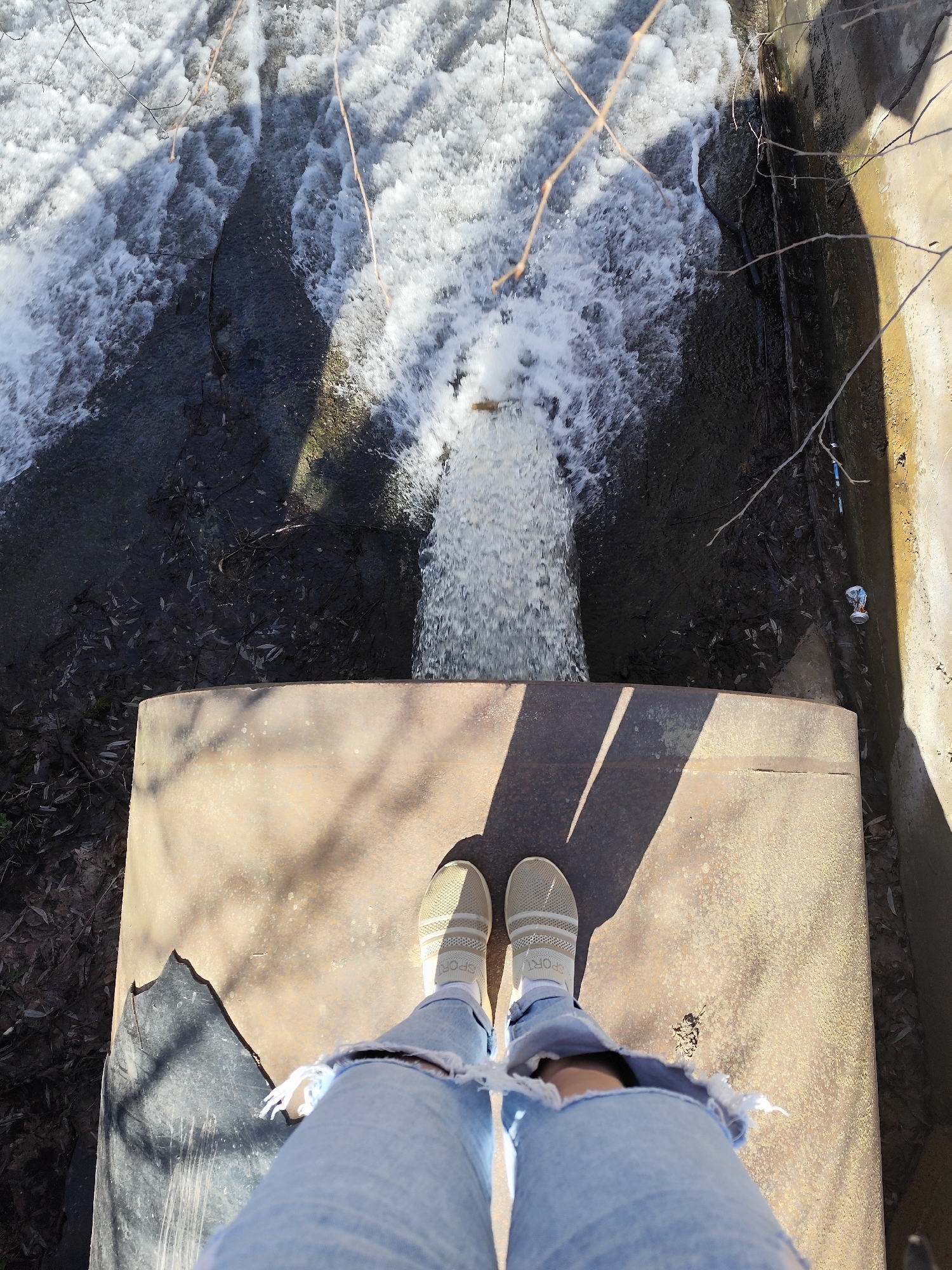 A high-angle shot depicts a person standing on a concrete structure above a fast-flowing body of water, likely a river or stream. The water appears turbulent, with white foam visible amidst darker currents.
The person is wearing light blue ripped jeans, white socks, and tan slip-on shoes with white soles and white stripes. The concrete platform on which they stand is weathered and stained, with shadows cast across its surface, suggesting a sunny day. There is damage on the platform, with the corner being ripped or broken away.
The surrounding environment includes vegetation and what appears to be a man-made channel containing the water. A discarded aluminum can is visible near the channel's edge.
The location is Фоки, Russia.