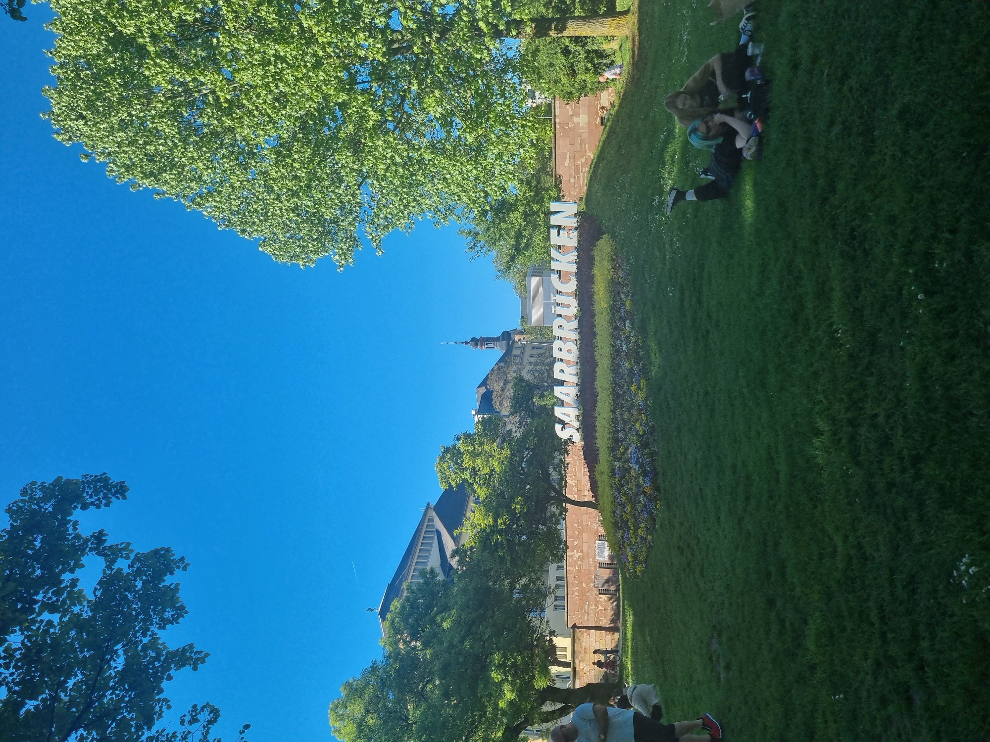 The image depicts a sunny scene in Saarbrücken, Germany. The prominent "SAARBRÜCKEN" sign is visible, indicating the location. A building with a tall spire is seen behind the sign. The scene includes a green lawn, trees, and clear blue sky. Several people are present in the image; one group is sitting or lying on the grass to the right, and another appears to be walking near the base of a tree.