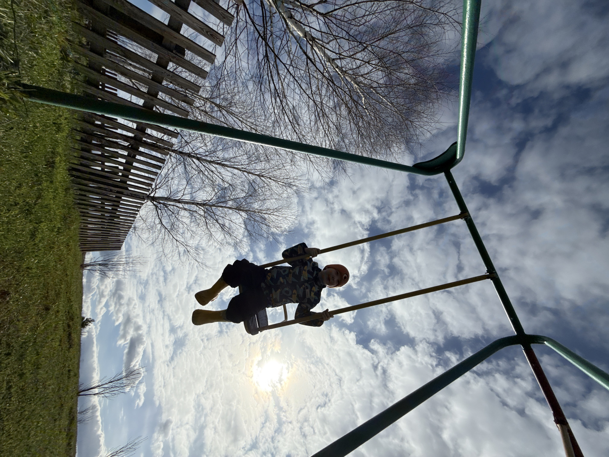 A low-angle photograph taken outdoors shows a child swinging on a swing set. The swing set frame is green and partially visible in the foreground and background. The child is wearing dark pants, a patterned shirt, a beanie, and light-colored boots. The child is facing the camera. The background contains a cloudy sky with the sun visible through the clouds, a bare tree, a wooden fence, and a patch of green grass. The photograph appears to have been taken in Nizhnyaya Chermoda, Russia.