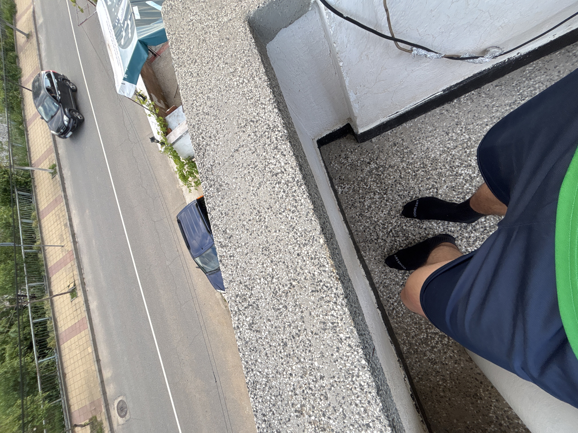 A high-angle shot depicts a person standing on a balcony, overlooking a street in Zlatograd, Bulgaria. The person is wearing dark socks and navy blue shorts with a green stripe. The street shows two cars parked and a road. The balcony is made of concrete. Above, exposed electrical wires are seen, some covered in aluminum foil.