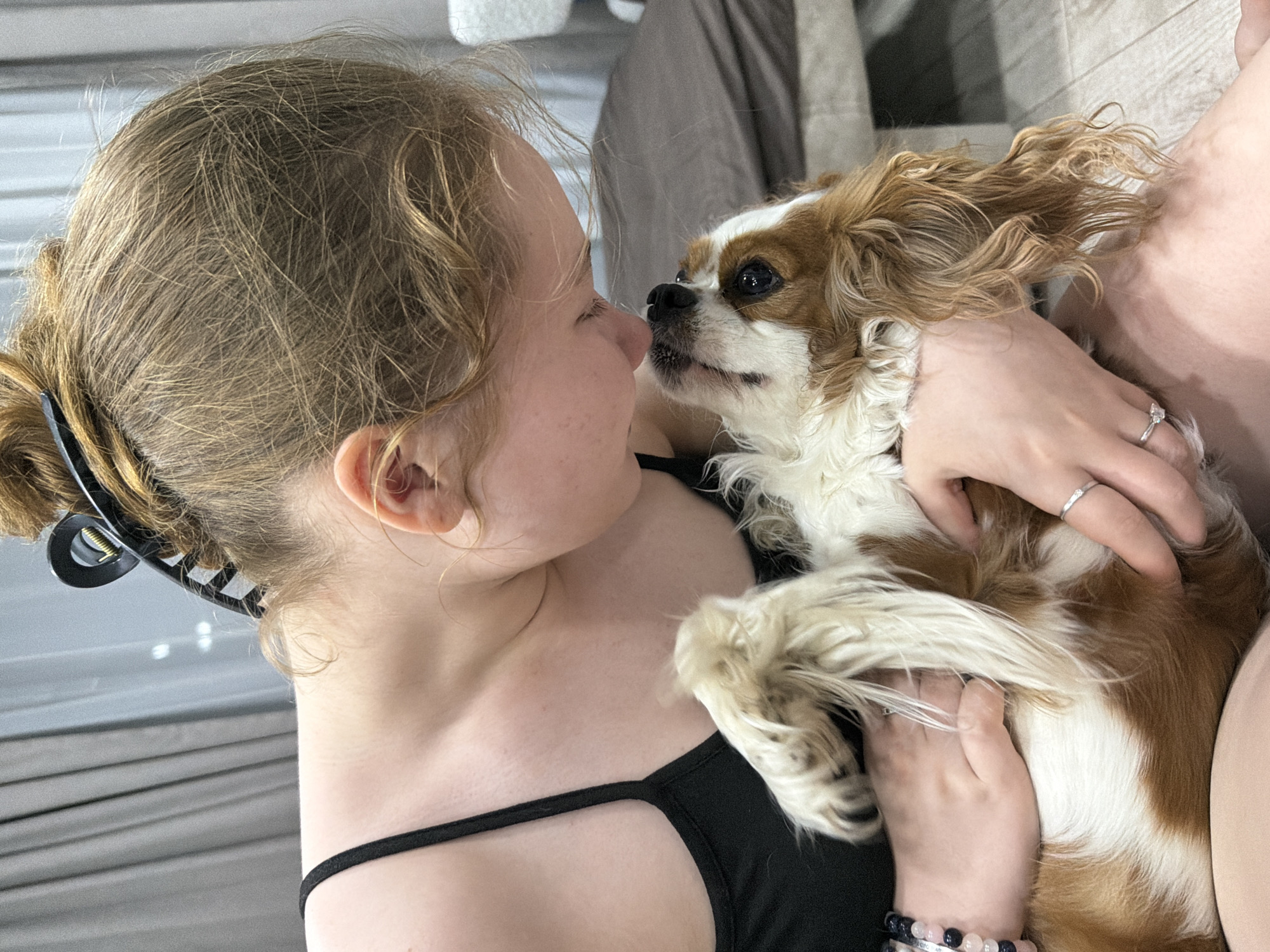 A woman with blonde hair pulled back with a black claw clip is holding a Cavalier King Charles Spaniel dog. The woman is wearing a black tank top and has rings on her fingers. She is looking at the dog, and their noses are almost touching. The dog is brown and white. The location is not specified. The city of Chaikovsky, Russia is not discernible in the image.