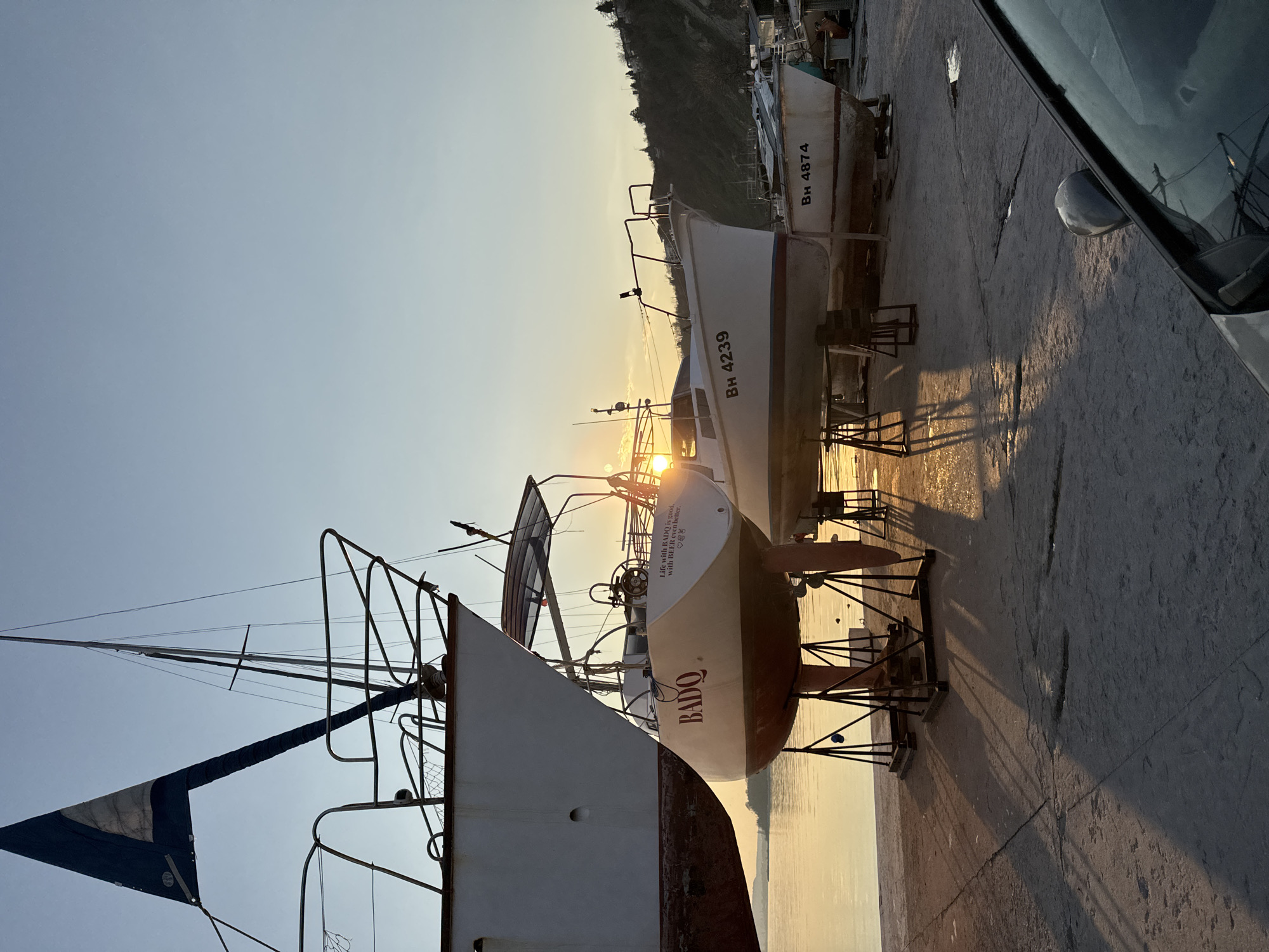 The image shows a row of boats on a concrete surface, likely a dock or marina area. The setting sun is visible in the background, casting long shadows. The boats appear to be of varying sizes and conditions, with some having visible names and numbers such as "BADO", "BH 4239" and "BH 4874". The presence of boats and the phrase "Life with BADO is good, with BEER even better" suggests a recreational or leisure-oriented setting. The background contains a treed hillside or bluff typical of coastal scenery. The photograph was taken in Varna, Bulgaria.