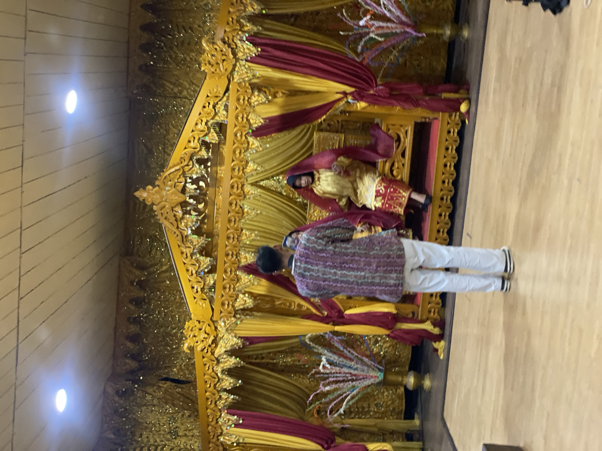 In Banjarmasin, Indonesia, a traditional wedding ceremony is underway. A woman, adorned in a gold dress and a red veil, is seated on a gilded throne, likely a bridal seat. A man, dressed in casual attire, stands facing her, his back to the camera. The backdrop is a cascade of gold and maroon curtains, with ornate golden carvings adorning the stage. The room's ceiling features wooden planks and recessed lighting, hinting at an indoor event. The floor is polished wood, reflecting the opulent decorations. The overall scene suggests a significant cultural event, possibly a wedding or a traditional reception.