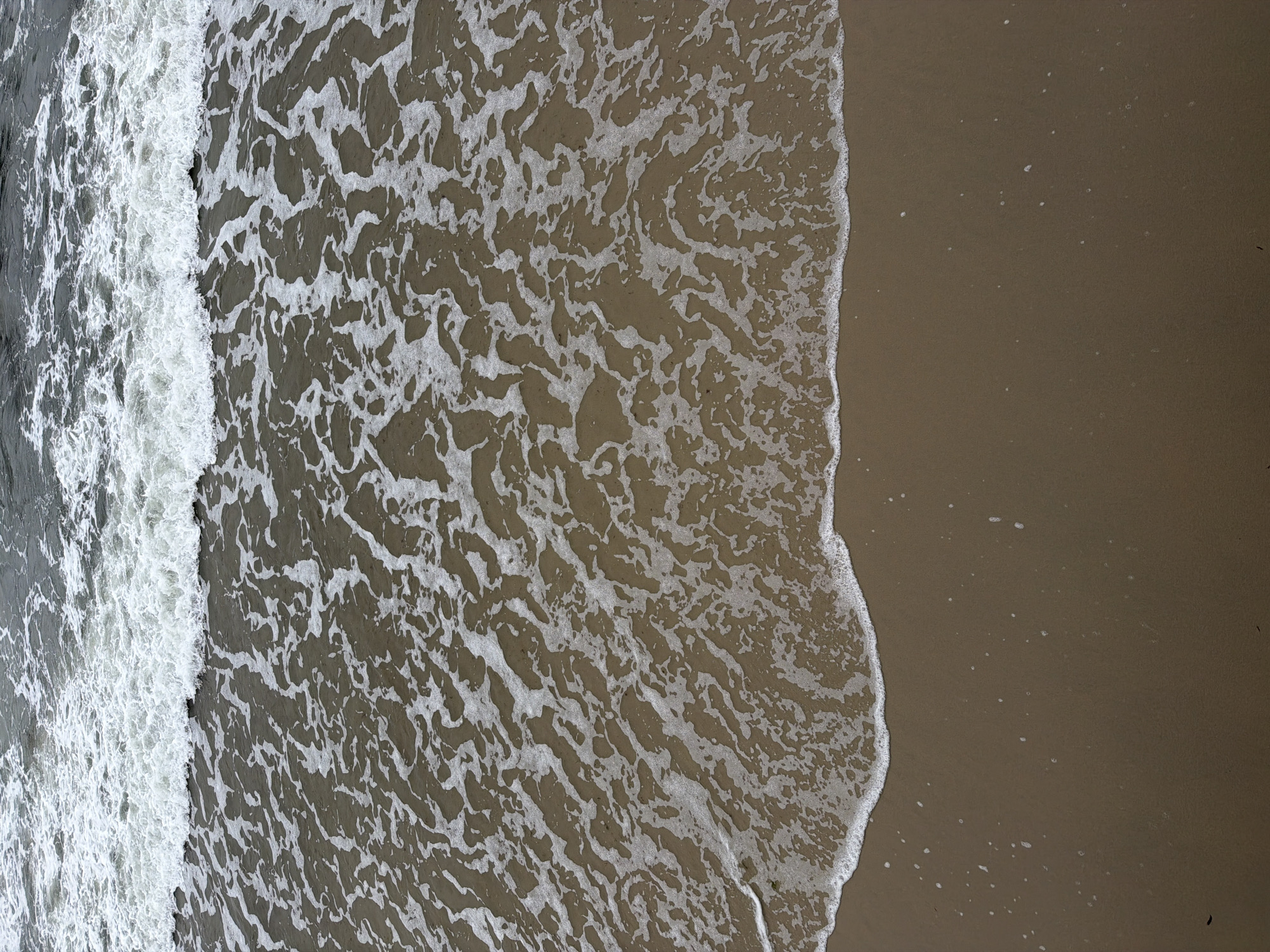 This is an aerial, full-frame shot of a sandy beach with waves coming in. The water is grey with white foam, and the sand is a light brown color. The scene appears to be a typical beach, devoid of any people or structures, and could be anywhere, including Zelenogradsk, Russia.