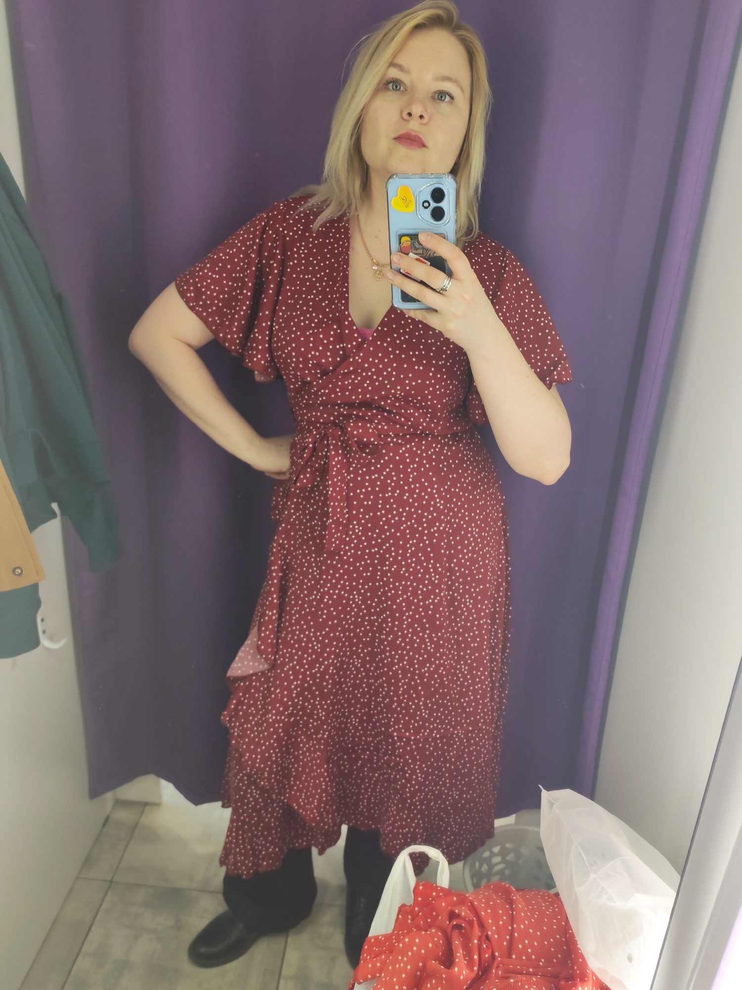 A blonde woman, appearing to be in her 30s or 40s, takes a mirror selfie in a retail changing room in Izhevsk, Russia. She stands facing forward, holding a light blue smartphone with a yellow heart sticker and other small stickers on its clear case, capturing her reflection. She wears a burgundy red wrap dress patterned with small white polka dots. The dress features short, ruffled flutter sleeves and a ruffled hemline, with a tie-waist detail. A pink bra strap is visible beneath the V-neck. She also wears black shoes or boots and a gold-colored necklace with a pendant. Her expression is neutral to slightly contemplative as she looks towards the phone.
The changing room has a deep purple curtain in the background and a light-colored tiled floor. To the left, a dark green coat and a lighter brown garment hang on a white hook against a white wall. On the floor to the right, a white plastic shopping bag contains a folded red garment, also with white polka dots, suggesting she is trying on different items. Another translucent white bag is partially visible further back. The scene depicts an indoor retail environment.