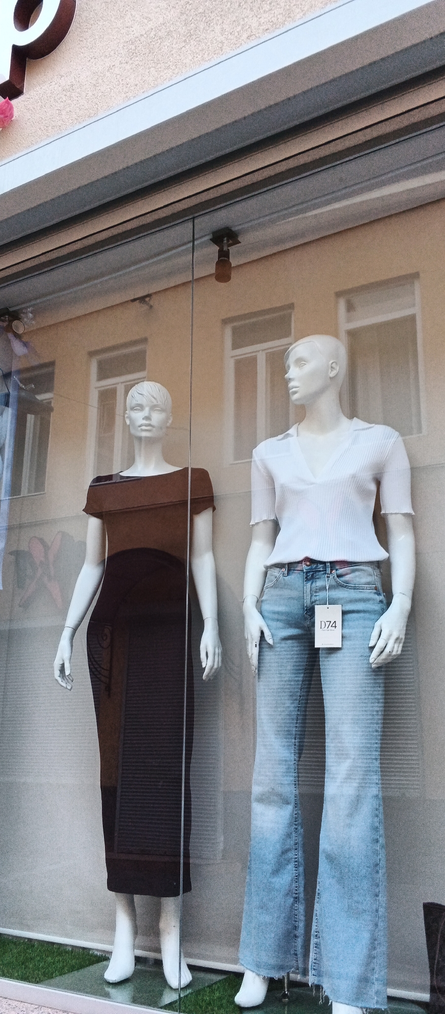 The image shows a shop window display featuring two mannequins. The mannequin on the left is wearing a long, dark brown dress with a textured pattern. The mannequin on the right is wearing a white, short-sleeved, collared shirt and light blue flared jeans with a price tag labeled "D74". Both mannequins are white and have stylized hair. They are positioned in front of a building with beige walls and windows. The display is set up on a surface covered with artificial green grass. The overall scene suggests a retail environment, likely a clothing store. There is no specific information in the image to place it in Mukachevo, Ukraine.