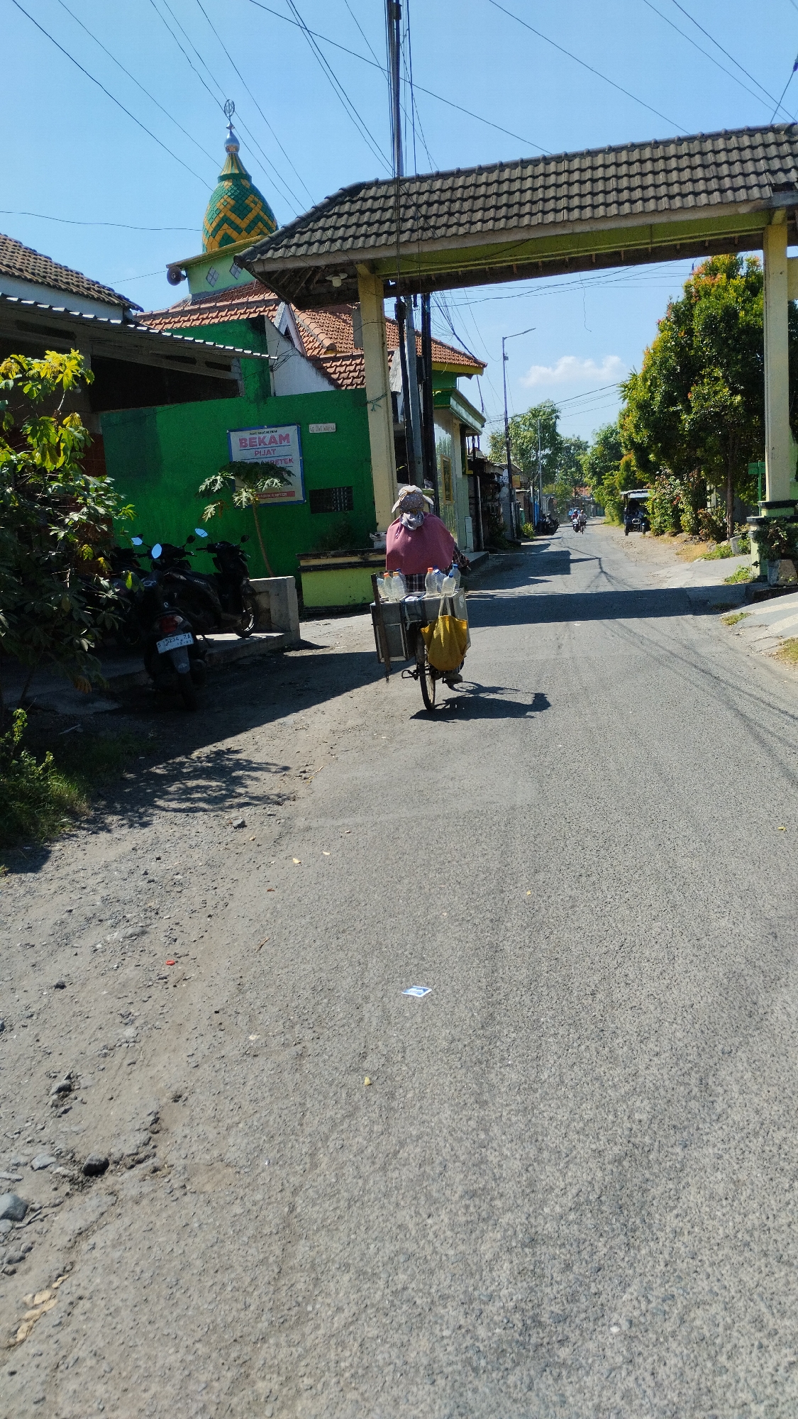 In Kramat Jegu, Indonesia, a woman is seen riding a bicycle equipped with a cart. She is wearing a sun hat and a pink garment covering her upper body, and a mask covering her lower face. Her cart carries several plastic bottles and a yellow bag. The bicycle is on a paved road, which appears to be a street lined with buildings and trees. To the left of the road, there is a green building with a sign that reads "BEKAM" and a mosque with a distinctive green and yellow dome. Several motorcycles are parked near the building. The sky is clear and blue, suggesting a sunny day. The road surface on the left is unpaved and rocky, contrasting with the paved road ahead. In the background, other vehicles and buildings are visible, indicating a populated area.