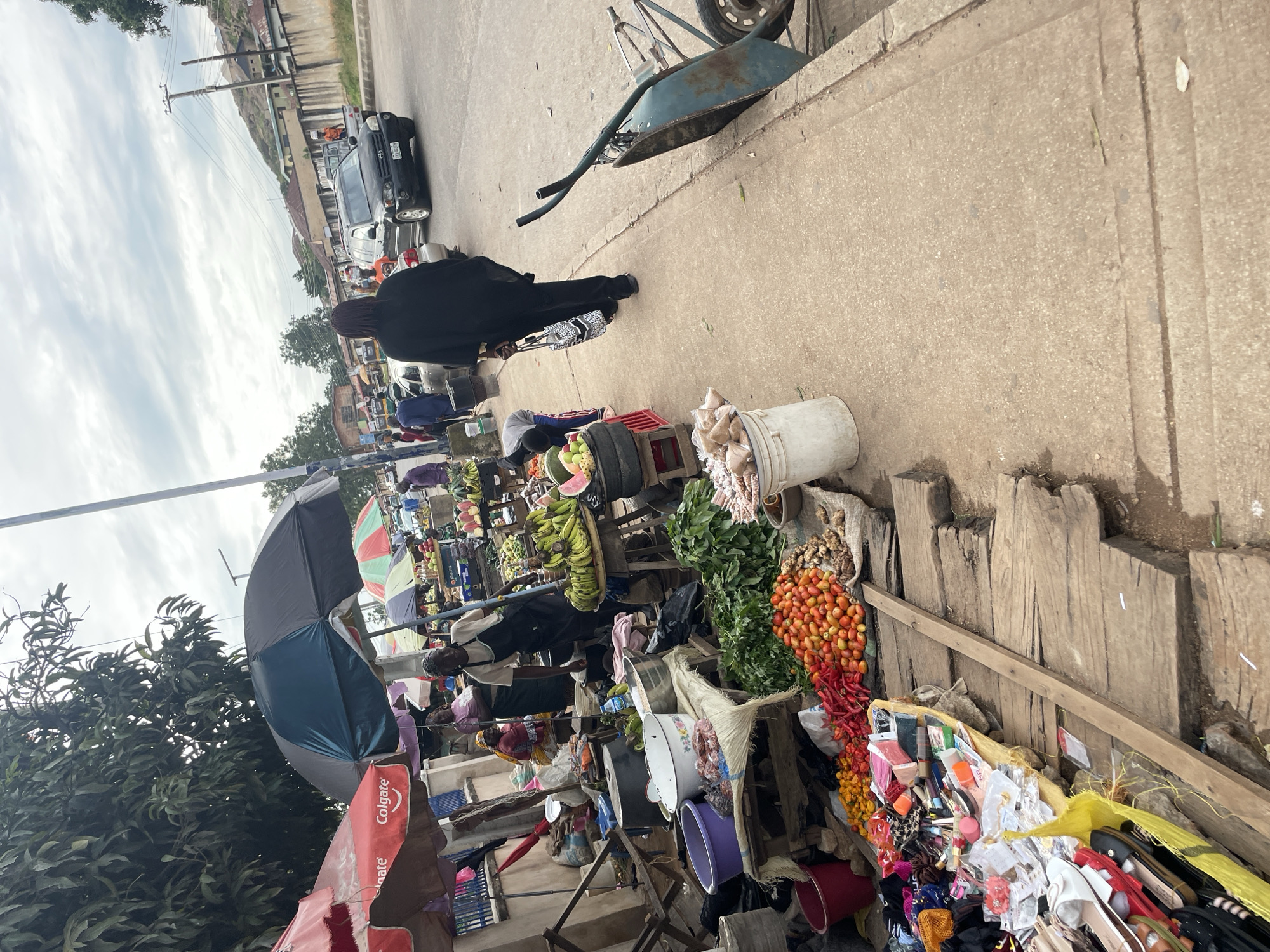 In Gura Top, Nigeria, a bustling street market lines the side of a paved road. Underneath large umbrellas, vendors display an array of fruits and vegetables. Bunches of bananas, tomatoes, and peppers are prominently featured. Various household items and clothing are also for sale. A woman in a black outfit walks down the sidewalk, carrying a patterned bag. A wheelbarrow rests by the roadside, and several cars and a truck are visible on the road, suggesting active commerce and transportation. The environment appears to be an overcast day, with a mix of buildings and vegetation in the background.