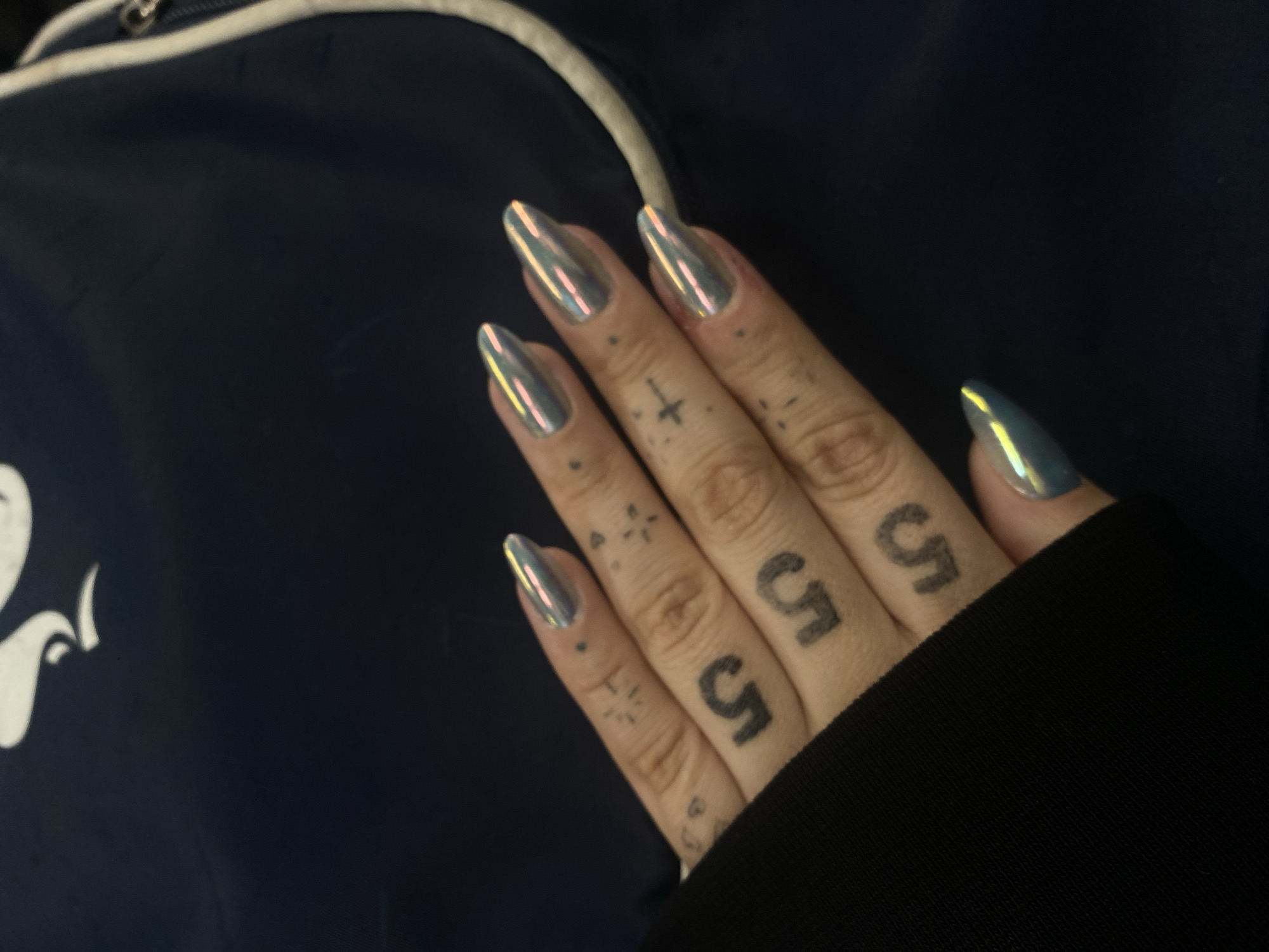 A close-up shot reveals a hand with metallic, pointed fingernails. The fingers are adorned with tattoos, including the number "5" on each finger and other small symbols. The hand is positioned against a blurred background of dark blue fabric with a white drawstring. Part of a black sleeve is visible in the lower right of the image. The location is unknown.