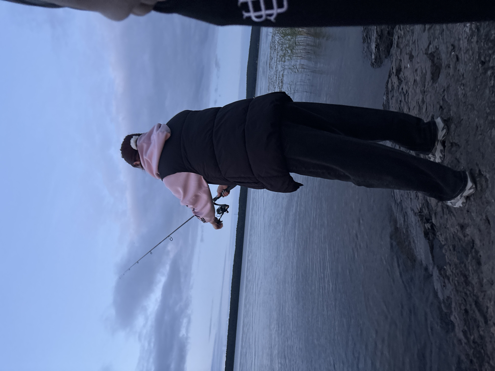 The image depicts a dimly lit, outdoor scene with a person standing on a rocky shore, fishing in a body of water. The person is seen from behind, wearing a black puffer vest over a pink hoodie and dark pants. They are holding a fishing rod, actively engaged in fishing. The water appears calm, reflecting the twilight sky which is predominantly blue with some clouds. In the distance, a dark line of trees borders the horizon. The overall atmosphere suggests a serene evening by the water. The location is identified as Shuiskaya Chupa, Russia, a place known for its natural beauty and lakes.