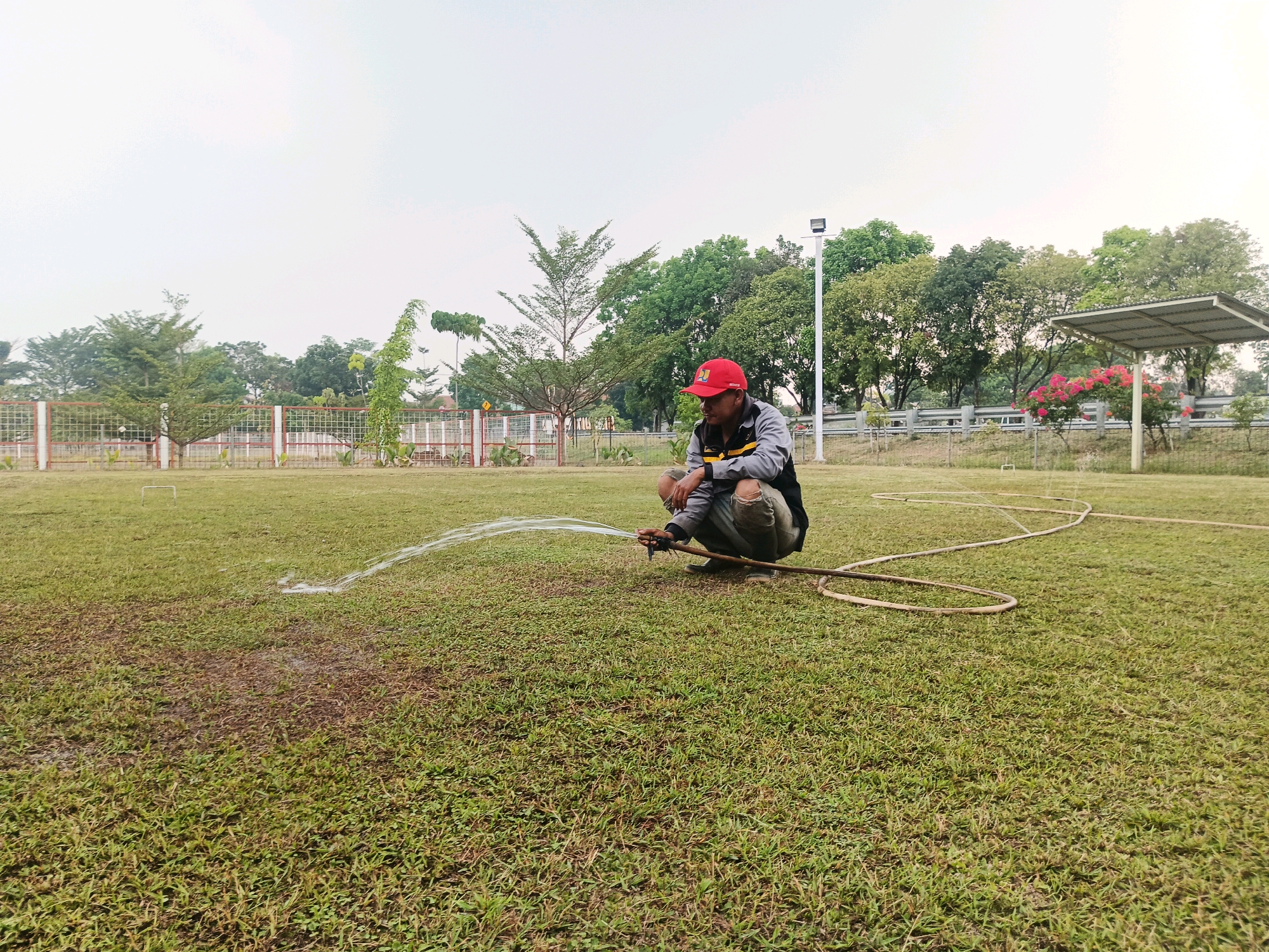 A man wearing a red cap and a grey and black jacket is squatting on a grassy field, holding a hose and watering the grass. The field appears to be a sports field with a faint outline of a goalpost visible in the distance. Trees and a fence line the background, with a covered seating area to the right. The sky is overcast. The scene is likely located in a park or recreational area within Bandung City, Indonesia.