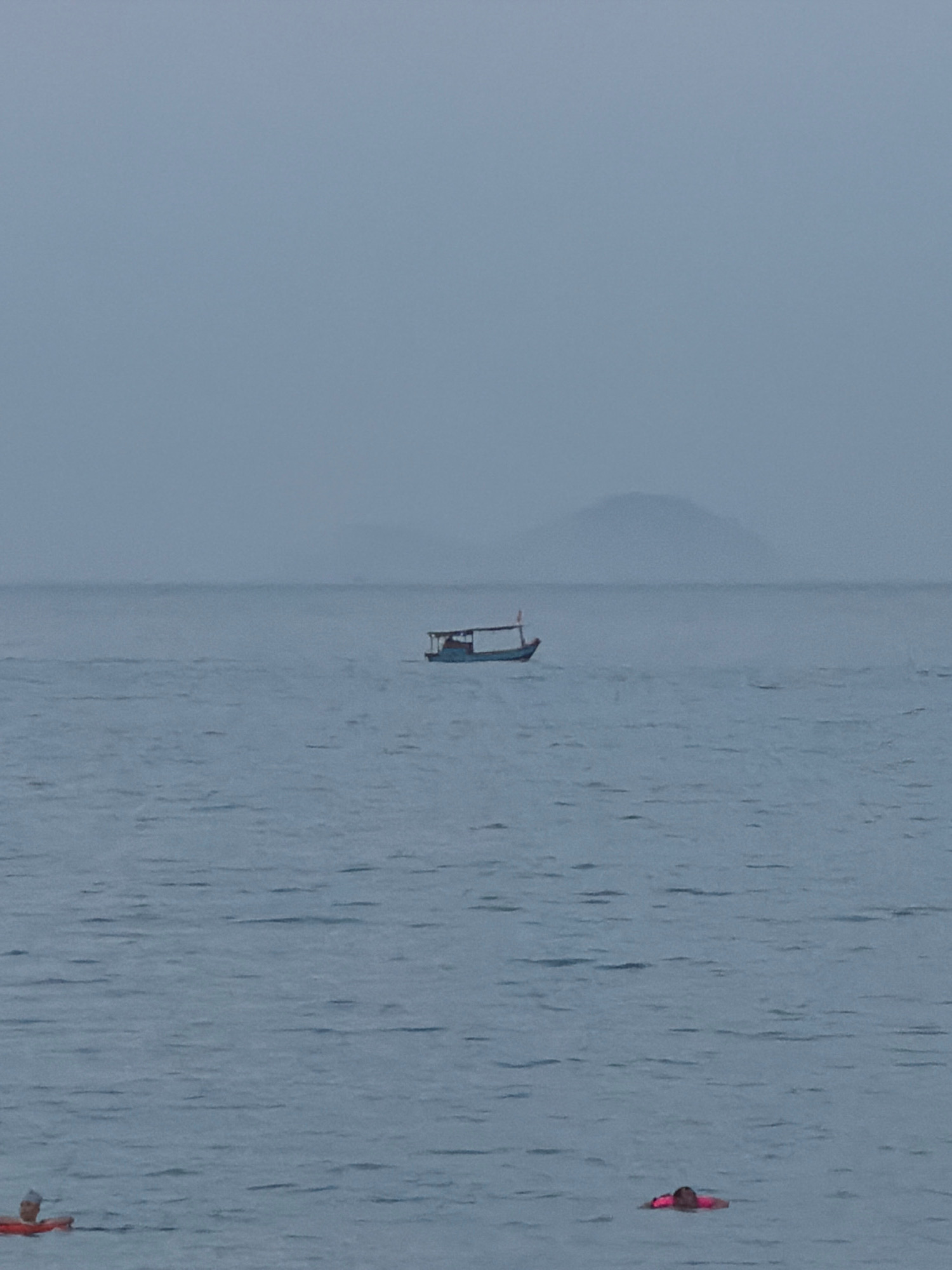 A lone fishing boat, painted blue with a canopy, navigates a calm, overcast sea. In the foreground, two individuals are visible in the water. One person, wearing a red floatation device, is partially submerged near the left edge of the frame. Further to the right, another person, wearing a pink life vest, floats on the surface of the water. Distant, hazy hills rise from the horizon. The scene appears to be taken from the shore of Nha Trang, Vietnam, during a muted, possibly foggy or overcast, day.