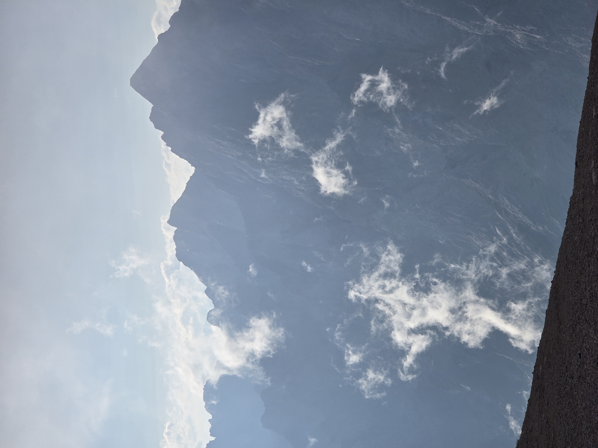 This image captures a majestic mountain landscape, likely in the vicinity of Garmisch-Partenkirchen, Germany, renowned for its alpine scenery. The scene is dominated by a series of rugged mountain peaks that recede into the distance, partially obscured by wisps of white clouds. The mountains are rendered in shades of blue and gray, with sharp ridges and dramatic slopes suggesting their formidable nature.
In the foreground, a dark, rocky slope extends diagonally across the bottom of the frame, providing a stark contrast to the softer textures of the mountains and sky. This foreground element adds depth and a sense of scale to the composition.
The sky above is a hazy blue, with scattered clouds that are illuminated by sunlight. Some of these clouds are close to the mountain peaks, giving the impression that they are clinging to the slopes, while others drift higher in the atmosphere. The overall lighting suggests it is either early morning or late afternoon, with a diffused quality that softens the harshness of the mountainous terrain. There are no visible people or man-made structures in the image, emphasizing the natural, untamed beauty of the mountain environment.