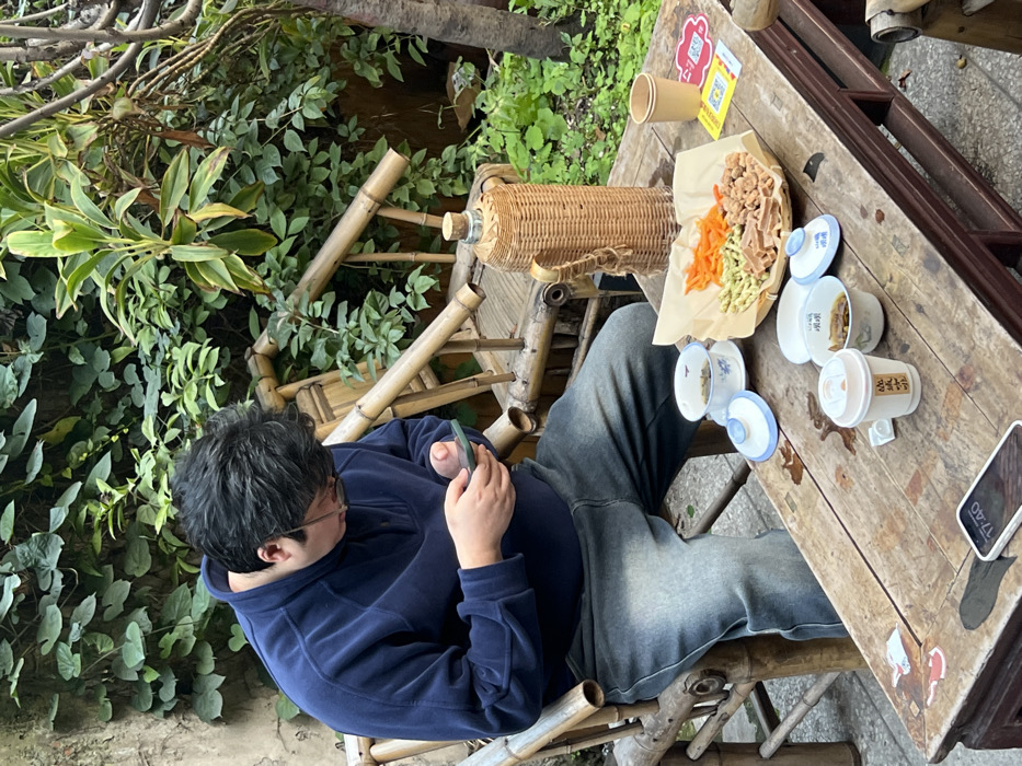 The image displays an outdoor setting in Yuzhong District, China, featuring a male individual seated at a rustic wooden table. The individual wears a dark blue fleece quarter-zip sweater and faded blue jeans, with dark hair and glasses. He is actively engaged with a smartphone, held in both hands, looking down at its screen. The phone has a green case.
The wooden table in front of him holds various items: a tall, wicker-wrapped thermos bottle with a cork stopper, and a plate lined with light brown paper presenting four distinct snacks—sliced orange carrots, green pea-shaped items, irregular brown crispy pieces, and rectangular light brown wafers. Also on the table are two small, white lidded teacups with blue patterns, one with its lid removed; two small, white lidded bowls with blue Chinese characters, one lid removed revealing a dark liquid or food; a white disposable coffee cup with Chinese text; and a plain brown paper cup. Another white smartphone, displaying "17:40", rests face-up on the table surface. A yellow QR code sticker is also visible on the table. The individual is seated on a bamboo chair. The background consists of dense green foliage, including vines on a wall and larger leafy plants, creating a natural, enclosed garden-like environment.