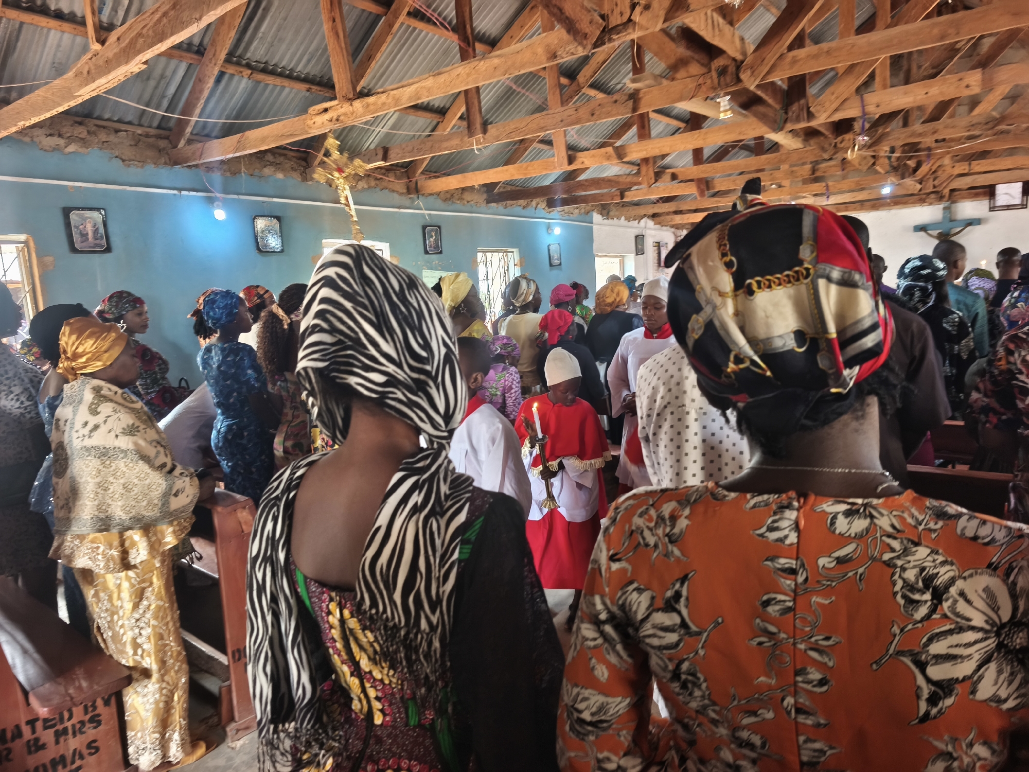 The image depicts a gathering of people inside a building that appears to be a church in Nabatum, Nigeria. The interior has a blue wall and a roof made of corrugated metal sheets supported by wooden beams. Religious pictures hang on the wall. The people are dressed in a variety of colorful traditional African attire, and many women wear head coverings. Some individuals are seated in pews while others stand. A person in a red robe holds a candle. The focus is primarily on the backs of people, suggesting the image was taken from behind the congregation.