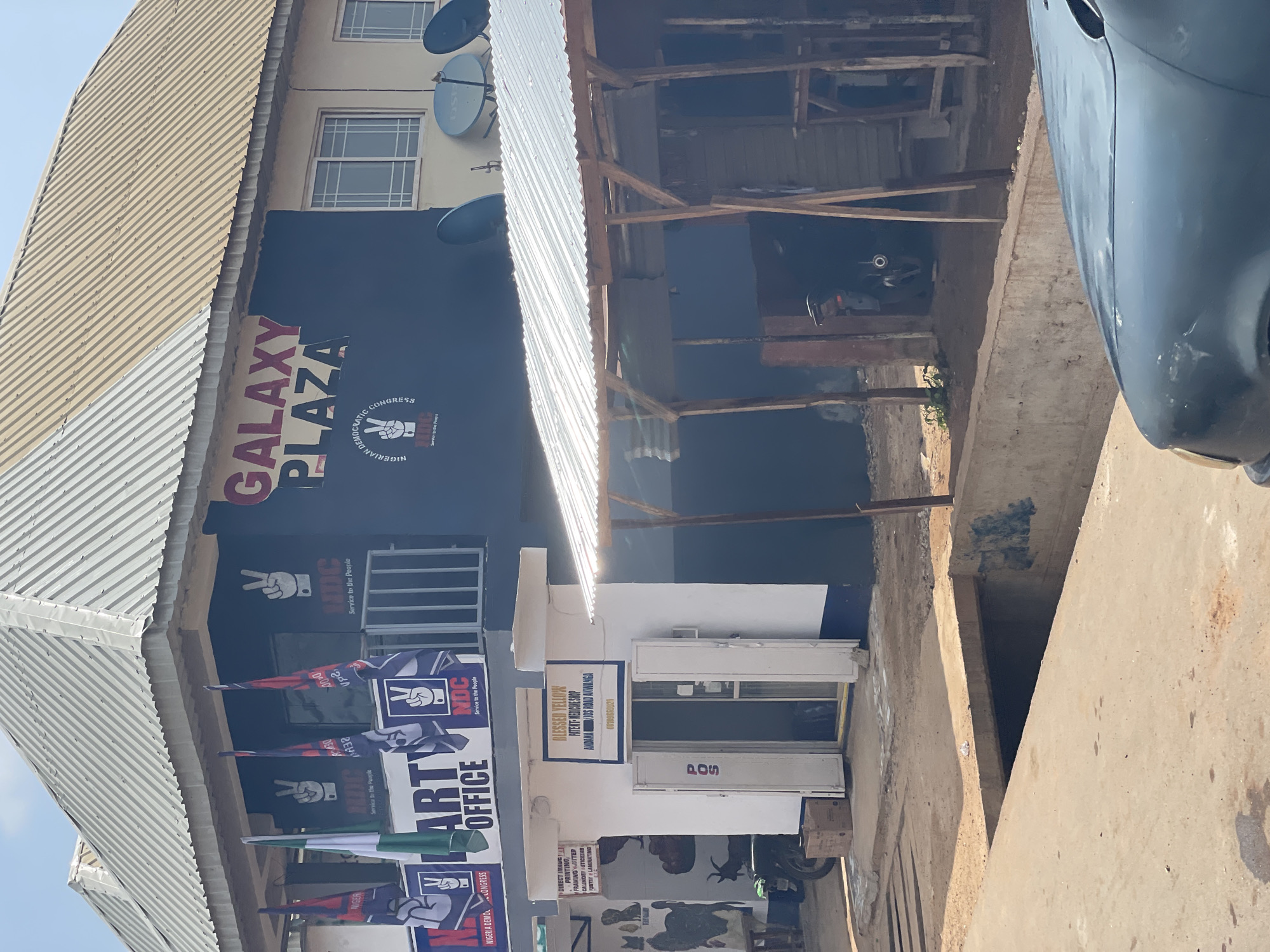 The image shows the front of the "GALAXY PLAZA" building, which houses the "NIGERIAN DEMOCRATIC CONGRESS" party office. Flags of the NDC party are displayed outside the building. A sign above a small storefront indicates "BLESSED YELLOW PATENT MEDICINE SHOP, ANDANA RD, UWADA AKWANGA." The building is located on a street with a paved road. A motorcycle is parked in front of the medicine shop. The overall scene depicts a commercial and political establishment in Akwanga, Nigeria.