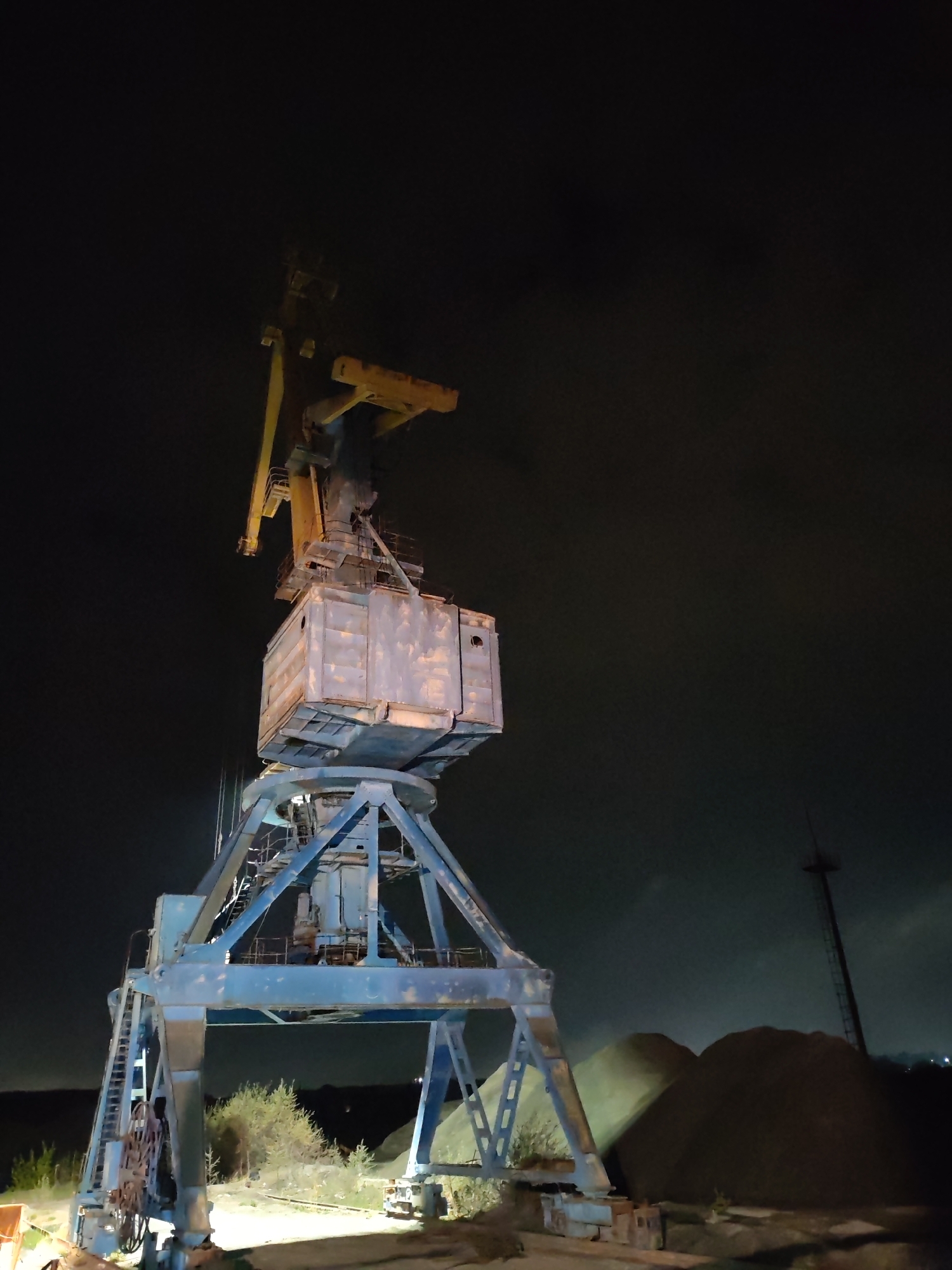 The photograph, taken at night, depicts a large, industrial crane located in what appears to be a construction or industrial area. The crane is predominantly blue and yellow, showing signs of aging and wear. It is composed of a tall supporting structure and a movable boom arm. The crane's base is situated near piles of dark material, possibly sand or gravel. In the background, there is a faint glow from city lights and what appears to be a tall, thin tower or mast. The sky is dark and obscured by clouds. The location is presumed to be near Tver, Russia based on the request. There are no people visible in the scene.