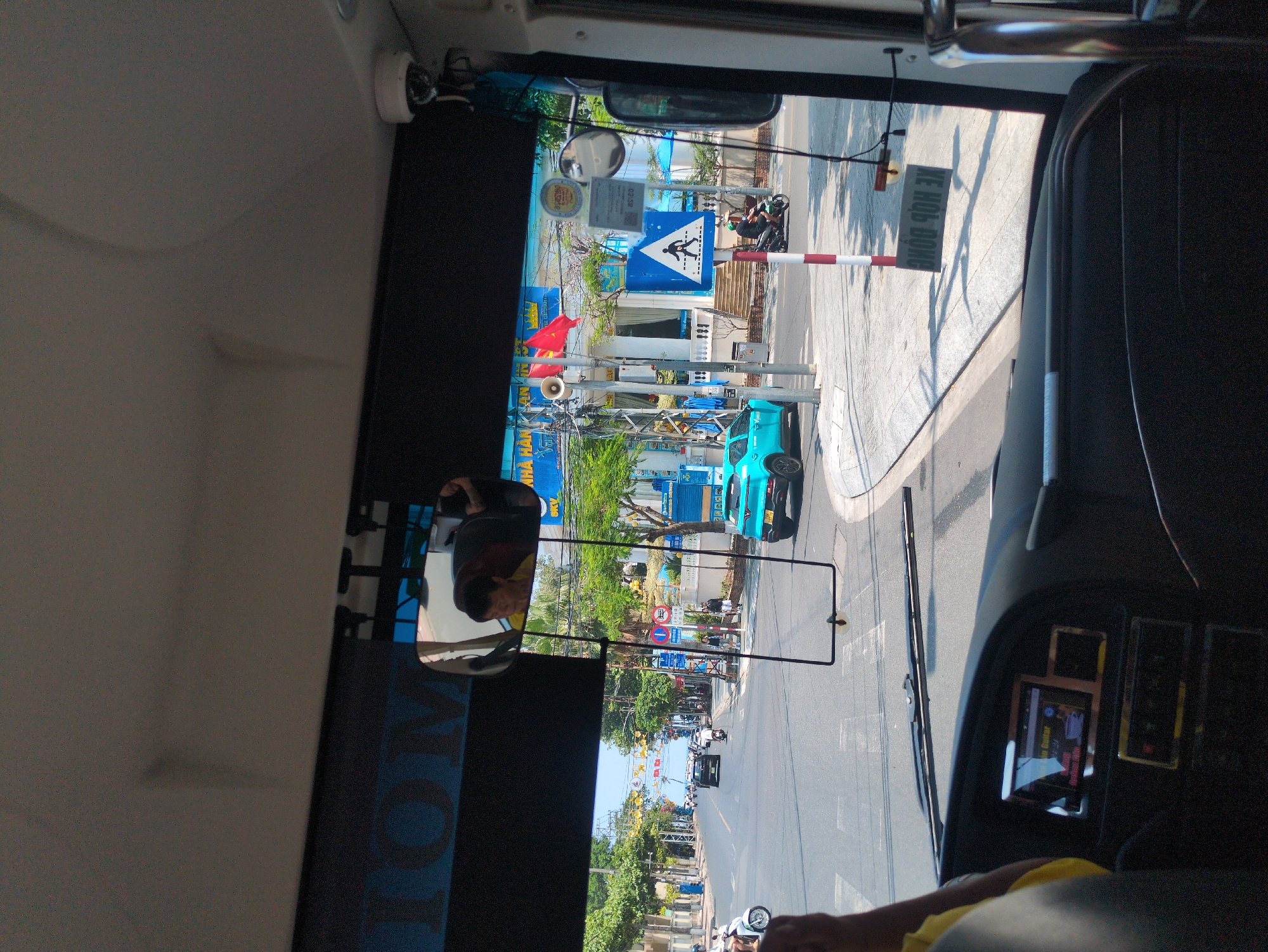 The image, taken from inside a bus, shows a street scene in Nha Trang, Vietnam. A blue car is parked on the side of the road, and pedestrians can be seen on the sidewalk. A pedestrian crossing sign is visible. The image also captures the bus's dashboard and rearview mirror, which reflects the driver. A sign is visible near the crosswalk reads "Xe Hop Dong".