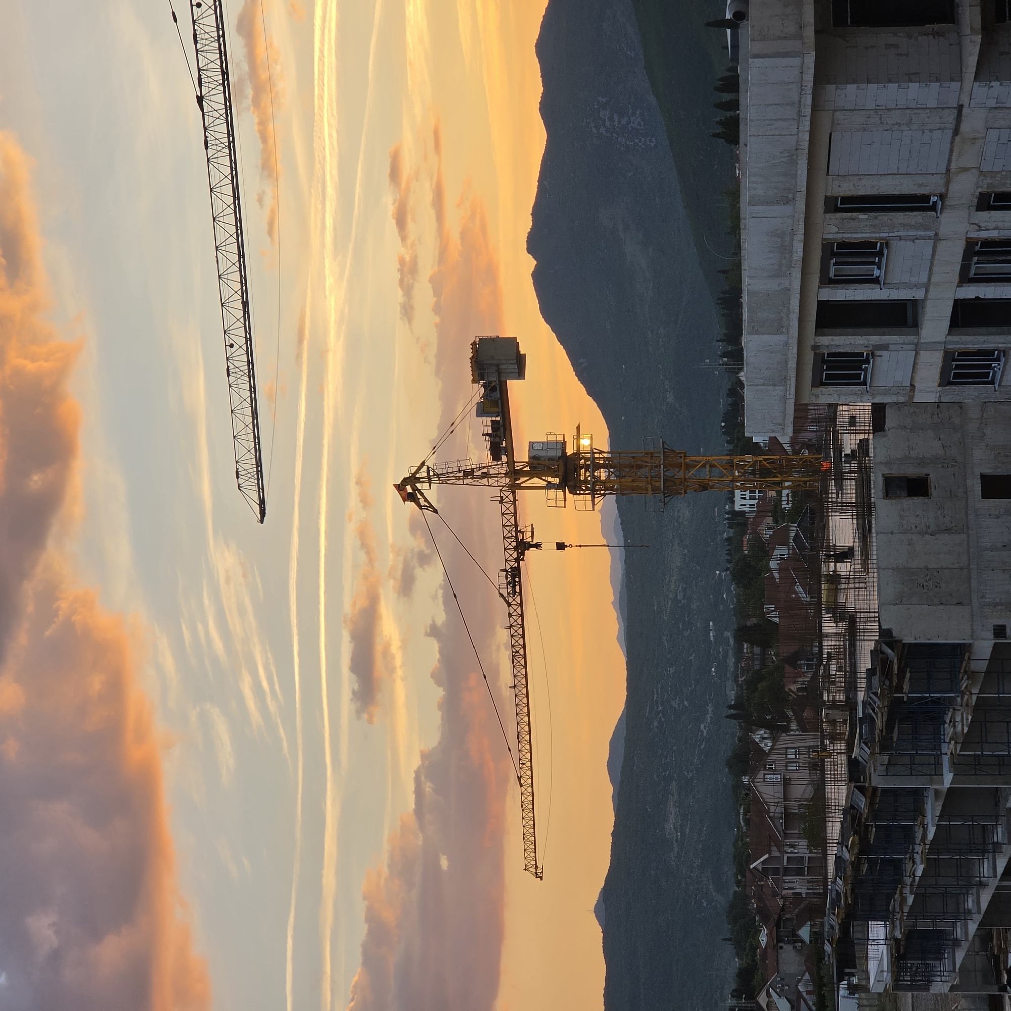 The image depicts a sunset view in Podgorica, Montenegro, featuring construction cranes against a backdrop of mountains and a building under construction. The sky is streaked with contrails and orange-hued clouds from the sunset. Two construction cranes are visible, one partially in the left upper corner and the other prominently in the center. A partially constructed building is on the right. The background shows a mountain range and the city of Podgorica nestled in the valley. No people are visible.
