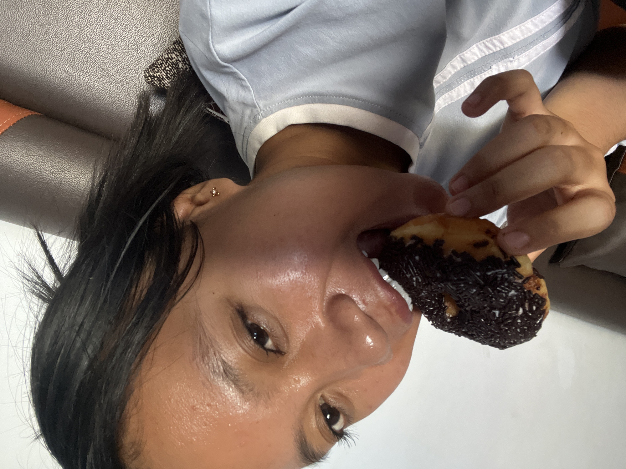 In an indoor setting, a young woman of Southeast Asian descent is captured in a close-up, head-and-shoulders shot, actively biting into a chocolate-frosted donut with dark sprinkles. She is wearing a light blue t-shirt with white piping around the collar and down the front. Her dark hair is visible, framing her face and falling over her shoulder. The woman's face is turned slightly towards the camera, with her right eye appearing more prominent. Her skin has a dewy sheen, possibly from sweat or product. A small, gold stud earring is visible in her right ear. The background consists of a textured, dark brown surface, likely upholstery, and a plain white wall. The scene does not provide any specific visual cues to pinpoint a location within the city of Jambearum, Indonesia.