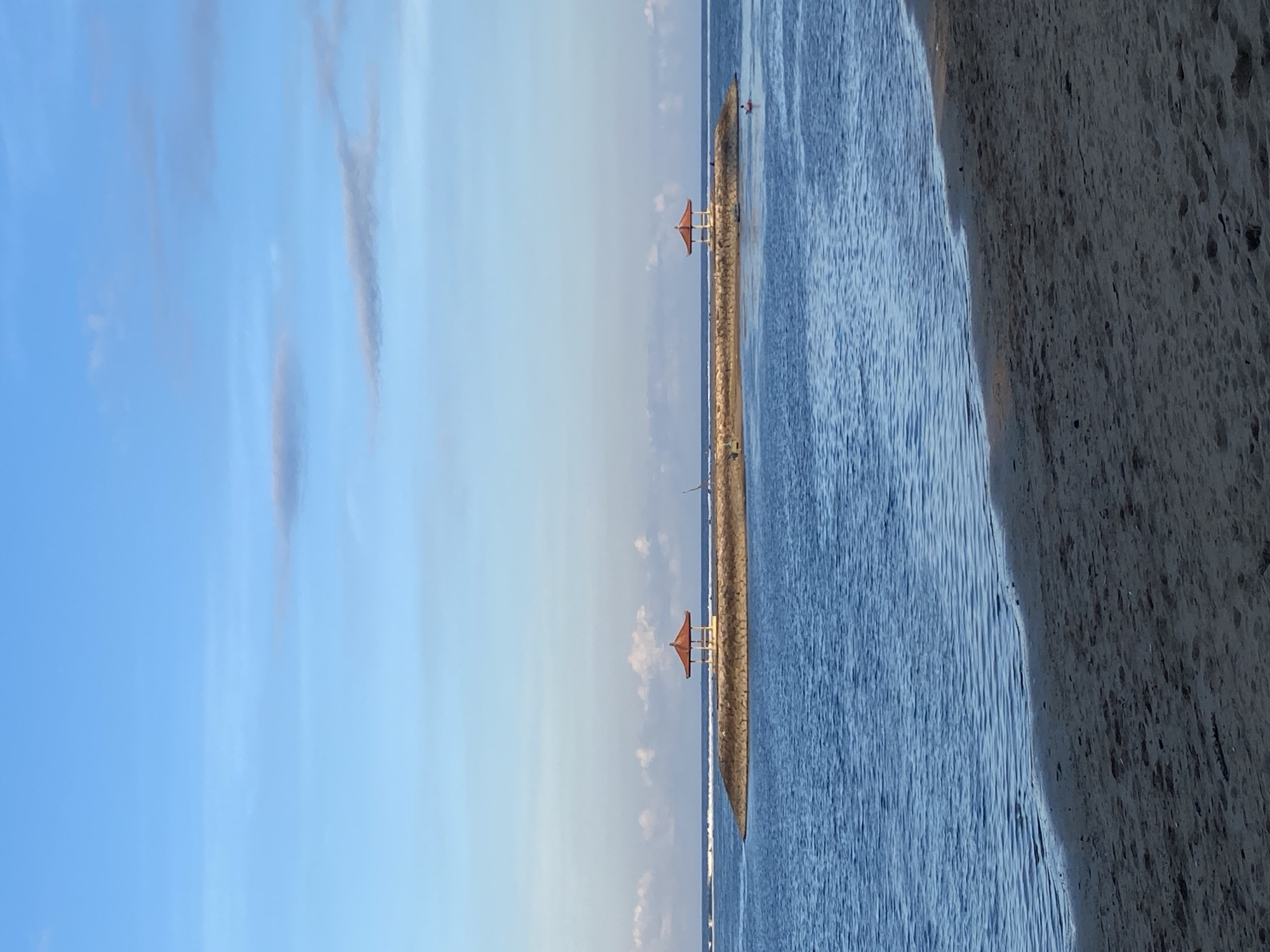 The image captures a serene beach scene at what appears to be dawn or dusk, given the soft light and gentle hues in the sky. The foreground shows a textured, sandy shore on the right, meeting a calm, deep blue ocean on the left. The water is slightly rippled, suggesting a peaceful atmosphere.
Stretching out into the sea is a man-made jetty, constructed from rough stones. On this jetty, a small, traditional-looking gazebo with a red roof and white pillars stands prominently. Further along the jetty, another similar structure can be seen, though it is partially obscured and less detailed.
The sky is a vast expanse of pale blue, transitioning to a lighter shade near the horizon, with wispy, faint clouds. The overall mood is tranquil and idyllic. In the far distance, on the water, a single person appears to be on a small boat or kayak, adding a subtle element of human presence to the otherwise natural landscape. There is no visible text in the image. The location context provided, Denpasar, Indonesia, suggests this is likely a coastal area within or near the city, known for its beautiful beaches and ocean views.