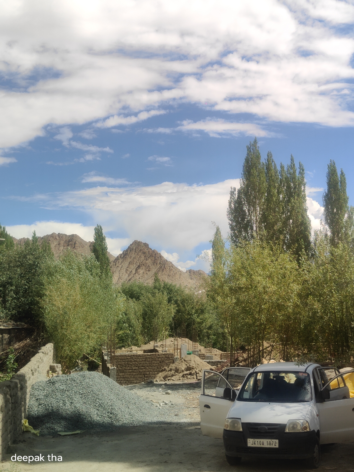 A white car with its doors open sits on a dusty road in front of a construction site in Leh, India. Piles of gravel and bricks are visible, along with unfinished brick walls. In the background, lush green trees and barren mountains under a cloudy blue sky complete the scene. The car has a license plate that reads "JK10A 1872." The text "deepak tha" is watermarked in the lower left corner of the image.