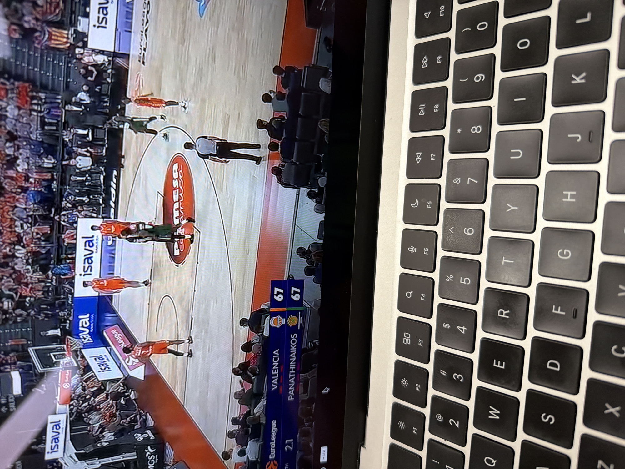 The image captures a laptop displaying a live basketball game on its screen, with the laptop's keyboard occupying the right half of the frame. On the screen, several basketball players are visible on the court; some wear orange uniforms, and others are in green. A referee stands near the center court. Spectators can be seen in the stands surrounding the court.
The activity taking place is the viewing of a live EuroLeague basketball match on the laptop. The scoreboard on the screen shows a tied game with both "VALENCIA" and "PANATHINAIKOS" having 67 points. The text "2.1" is visible below "EuroLeague", likely indicating the quarter or time remaining. The word "LIVE" confirms it's a real-time broadcast.
Notable details include the presence of sponsor logos like "isaval" prominently displayed on the court and advertising boards. The image is taken from a slightly elevated, oblique angle, showing the top-down view of the laptop's keyboard. The keyboard appears to be a MacBook or similar, featuring standard QWERTY layout keys, including function keys (F1-F10) with symbols for screen brightness, Mission Control, media playback controls, and volume. The photo itself was taken in New York, United States of America.
Visible text on the screen includes "isaval" (multiple instances), "EuroLeague", "2.1", "VALENCIA", "67", "PANATHINAIKOS", "67", "LIVE", "MRASA", and "aldin". The keyboard displays all standard alphanumeric characters, function symbols, and modifier keys like Shift, Command, and Option.