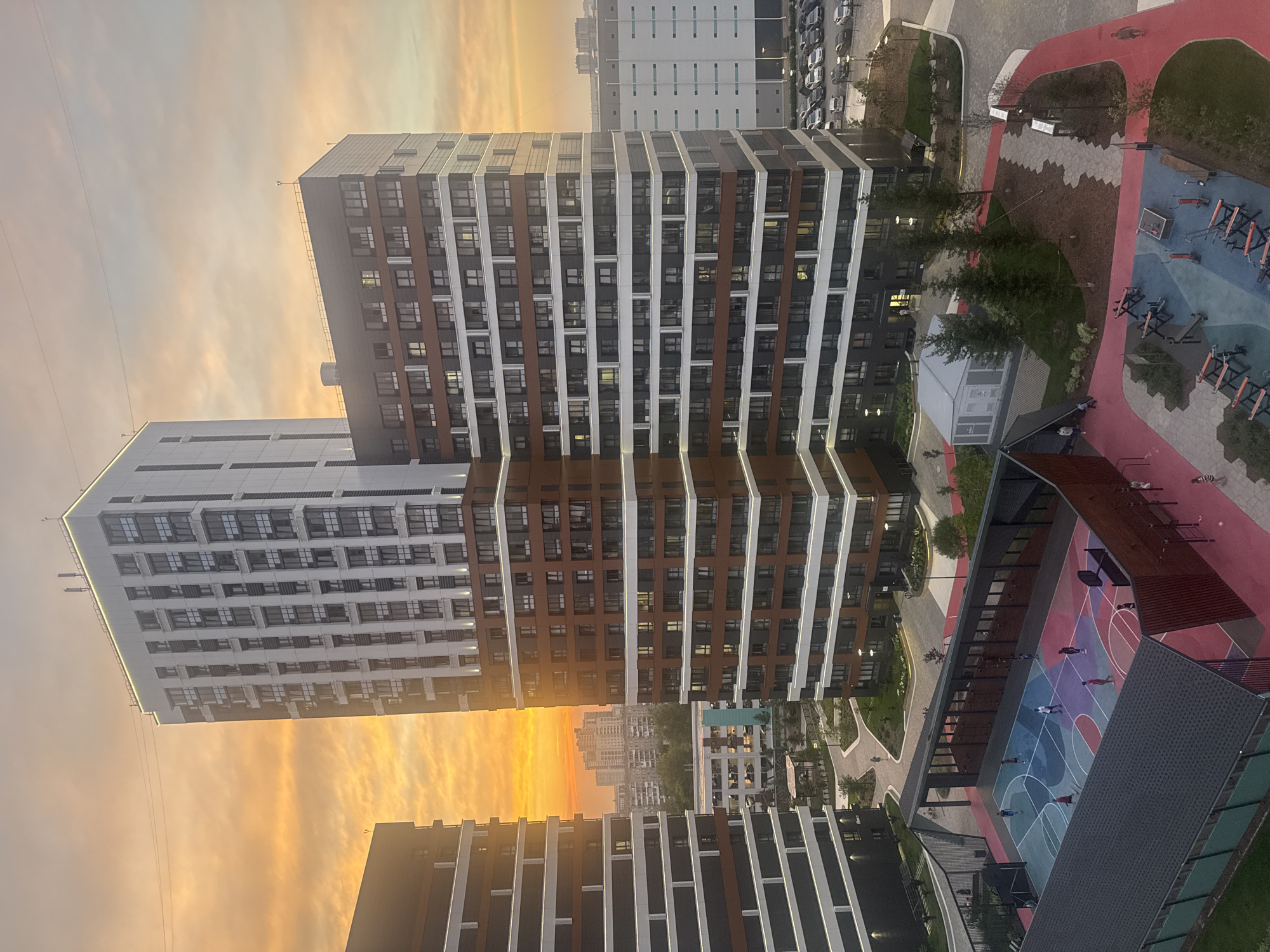 The image captures a modern residential complex in Novosibirsk, Russia, bathed in the warm hues of a sunset. Tall, multi-story buildings with dark brown and white facades dominate the skyline. One particularly tall building, clad in white with dark horizontal accents and numerous windows, stands prominently. Adjacent to it, another building features a darker facade with repeating patterns of windows and reddish-brown horizontal banding.
In the foreground, a vibrant, multi-colored outdoor recreational area is visible. This includes a basketball court with various colored sections, surrounded by a covered seating area. Further out, an open-air gym with multiple workout machines is set up on a blue and red tiled surface, bordered by red pathways. Several individuals are scattered throughout this area, some engaged in activities on the basketball court and others walking or interacting in the vicinity of the gym.
The background reveals more urban elements, including additional buildings of varying heights and a parking lot filled with cars. The sky above is a dramatic canvas of orange and yellow, indicative of a setting sun, with wispy clouds adding texture to the scene. The overall environment suggests a newly developed urban residential zone, designed with amenities for its residents.