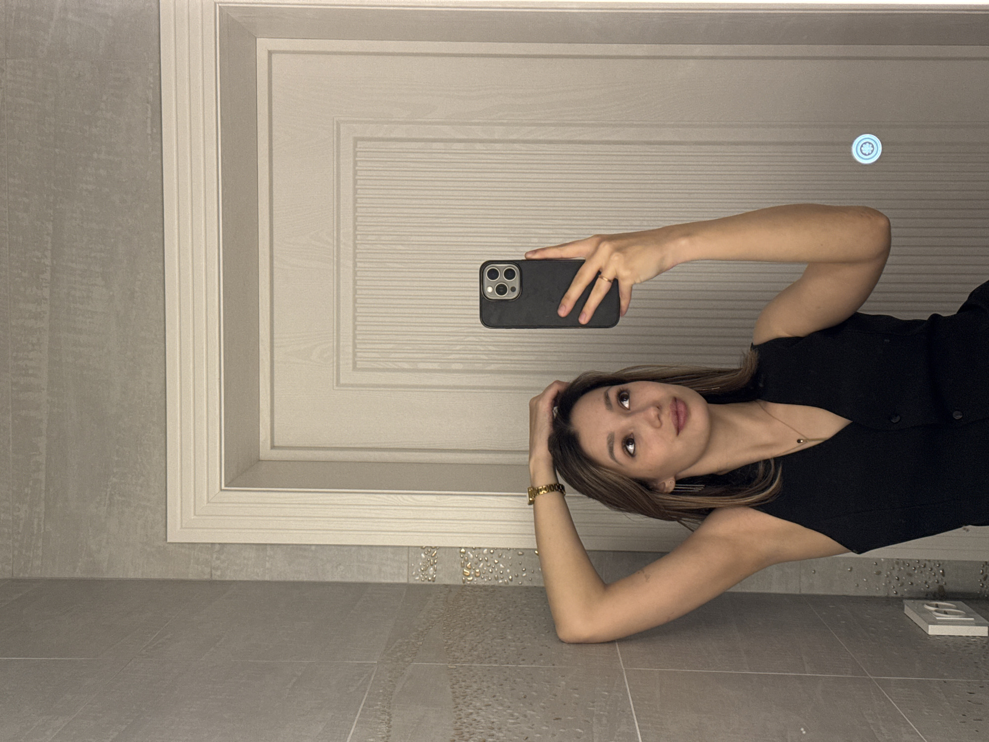 The image, rotated 90 degrees counter-clockwise, features a young woman taking a mirror selfie. She is depicted as lying on her left side on a light grey tiled floor. Her left arm is bent, with her hand resting near her head, while her right arm holds a dark-cased smartphone directly in front of her, facing the viewer. She gazes towards the phone with a calm, slightly pensive expression.
The woman has shoulder-length, light brown hair and is dressed in a black sleeveless top or vest. She wears a delicate gold necklace with a small pendant, a simple gold ring on her right hand, and a gold-toned watch on her left wrist.
The background reflects an indoor setting, likely a modern bathroom or dressing area. A white paneled door with intricate horizontal slat detailing is prominent, framed by a light grey tiled wall. A decorative strip of smaller, pebble-like textured tiles runs along the bottom of the wall, meeting the larger light grey floor tiles. In the upper right section of the image (as presented), a small, circular, illuminated fixture, possibly a light or vent, is visible on the door's reflection. An electrical outlet is noticeable on the tiled floor in the lower right corner. The lighting appears to be ambient interior light. There is no visible text in the image.