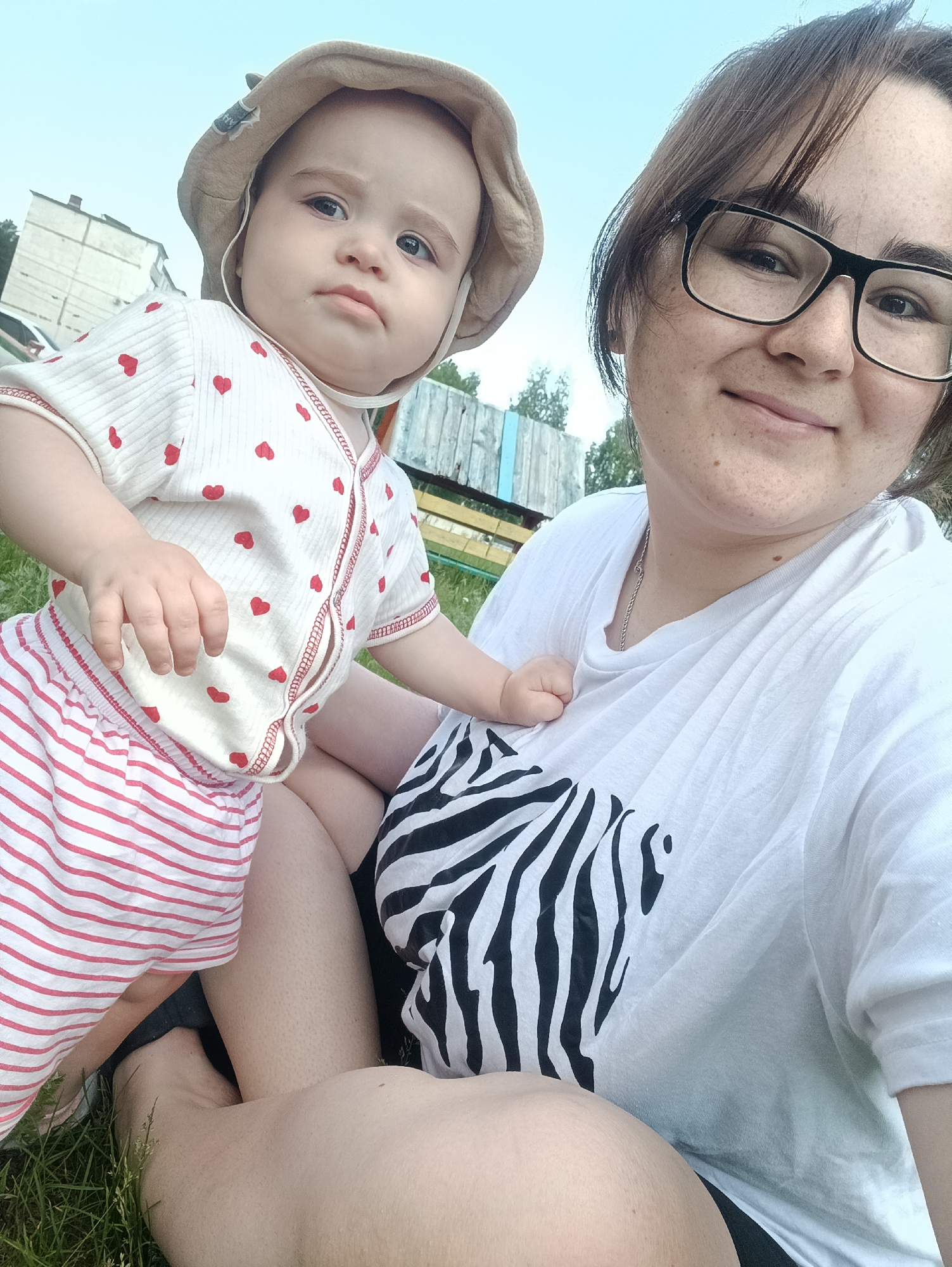 In the city of Noviy, Russia, a selfie is taken outdoors on a grassy area with a wooden fence and a building in the background. A young woman with glasses and brown hair is smiling at the camera. She is wearing a white t-shirt with a black and white zebra print on the chest. Next to her, a baby is sitting, looking at the camera with a serious expression. The baby is wearing a light-colored bucket hat and a white onesie with red hearts. The baby is also wearing striped pink and white shorts. The woman's right arm is around the baby, and the baby's left hand is resting on the woman's chest. The ground is covered with green grass.