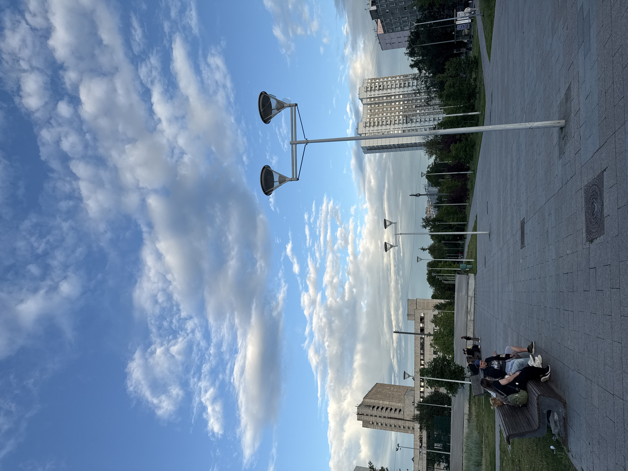 The image captures an outdoor scene, likely in Moscow, Russia, during the day. The sky is a vibrant blue, filled with scattered white clouds, suggesting fair weather. The foreground features a paved walkway with a patterned surface made of large gray tiles. Several modern lampposts with a distinctive dual-head design are spaced along the walkway, their metallic poles reflecting the light.
In the middle ground, a park-like area with green grass and trees is visible. Beyond the greenery, several multi-story buildings, typical of urban architecture, can be seen. One prominent building is a tall, light-colored structure with many windows, appearing to be an apartment building or office block. Another is a more Brutalist-style concrete building.
In the foreground on the right side, a group of people are resting on a park bench. One person is wearing a black hoodie and a blue baseball cap, facing towards the other individuals. Another person is wearing a black top and a green backpack. Their postures suggest a relaxed atmosphere. Further in the background, more people are seen walking or standing near the buildings. The time of day appears to be late afternoon or early evening, given the angle of the light and the slight dimming of the sky. There is no visible text in the image.