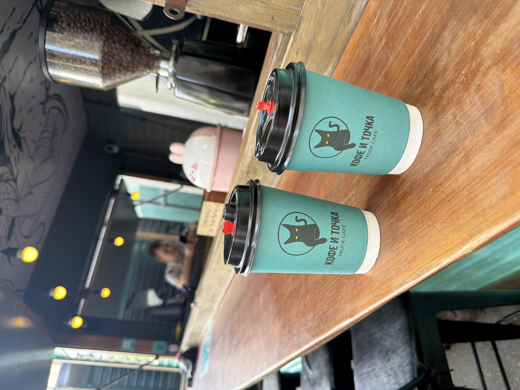Two paper coffee cups sit on a wooden countertop. The cups are teal with black lids and red stoppers. A logo of a black cat and the words "КОФЕ И ТОЧКА TRUCK CAFE" are printed on the cups. Behind the cups are coffee beans in a glass container, a pink bunny-shaped object, and a reflective surface that shows a person. The setting appears to be a coffee truck. The specific location is the city of Bac Nha Trang Ward, Vietnam, implied from the cafe's name (possibly Russian-owned or inspired).
