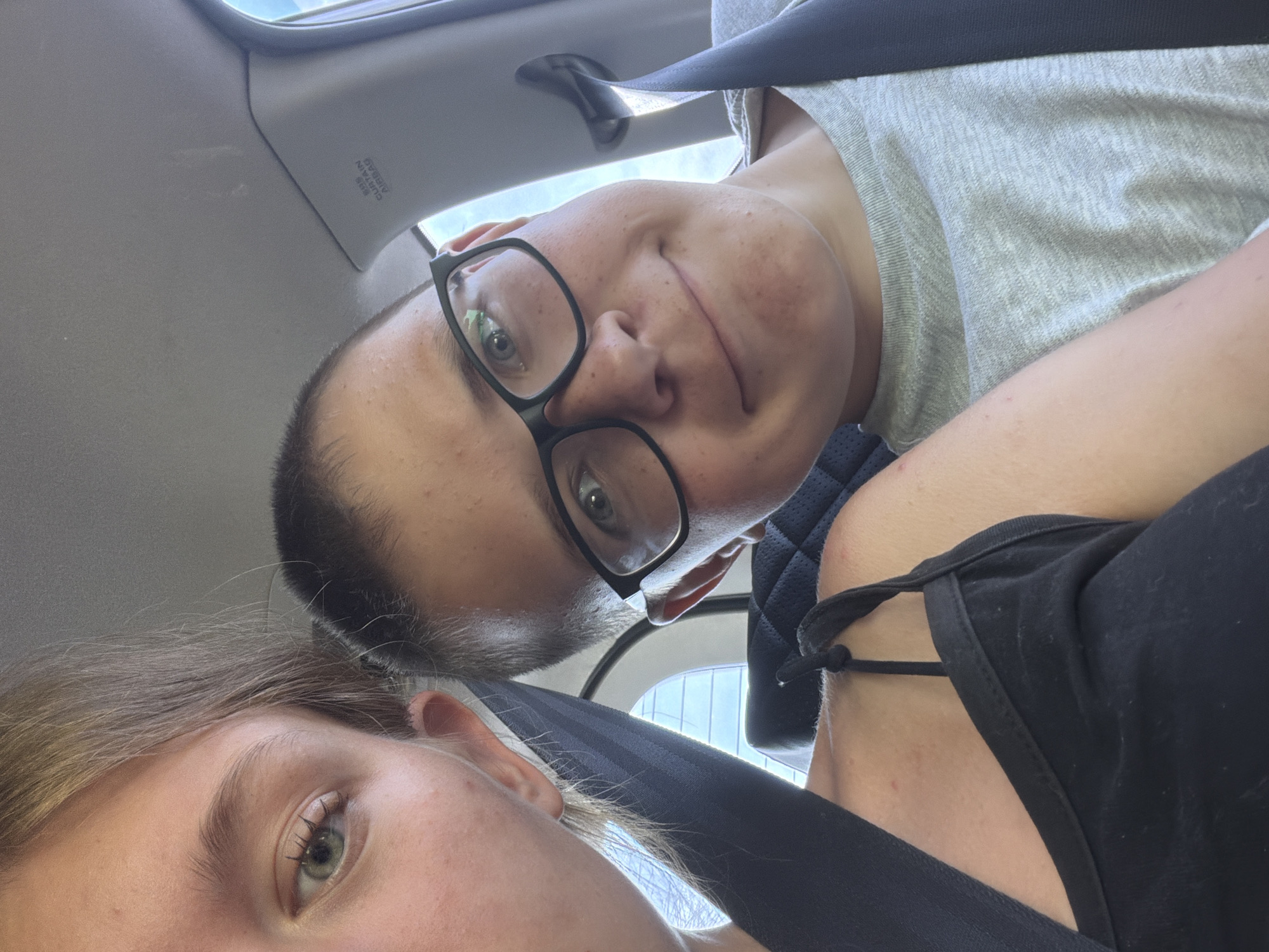 This is a selfie taken inside a car. Two young people are visible. On the left, a person with blonde hair and a green eye is partially seen, with their head and shoulder in the frame. They are wearing a black top with thin straps. On the right, a young person with short brown hair and glasses is in focus. They are wearing a grey t-shirt and smiling. Both individuals are wearing seatbelts. The background shows the car's interior, including a headrest, a seatbelt, and a portion of the car's roof with an "SRS CURTAIN AIRBAG" label. The overall lighting suggests it is daytime. There is no discernible information in the image to indicate a specific location within the city of Proleika, Russia.