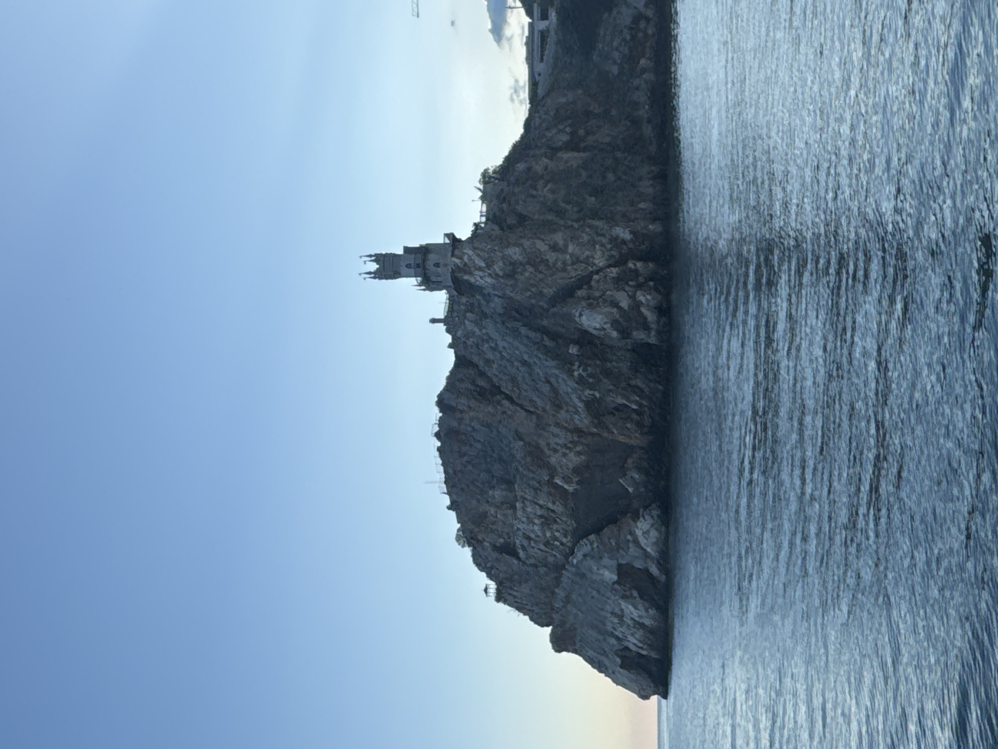 The image depicts Swallow's Nest, a decorative castle located on a cliff near Gaspra, in the Гаспринский поселковый совет region of Ukraine. The castle sits atop a rocky promontory. The sea surrounds the base of the cliff. The sky is a clear, pale blue. The photo is taken from a low angle, with the castle appearing vertically oriented. The castle's architectural details are visible, with its spires and turrets. The sea displays vertical lines, suggesting ripples on the surface.