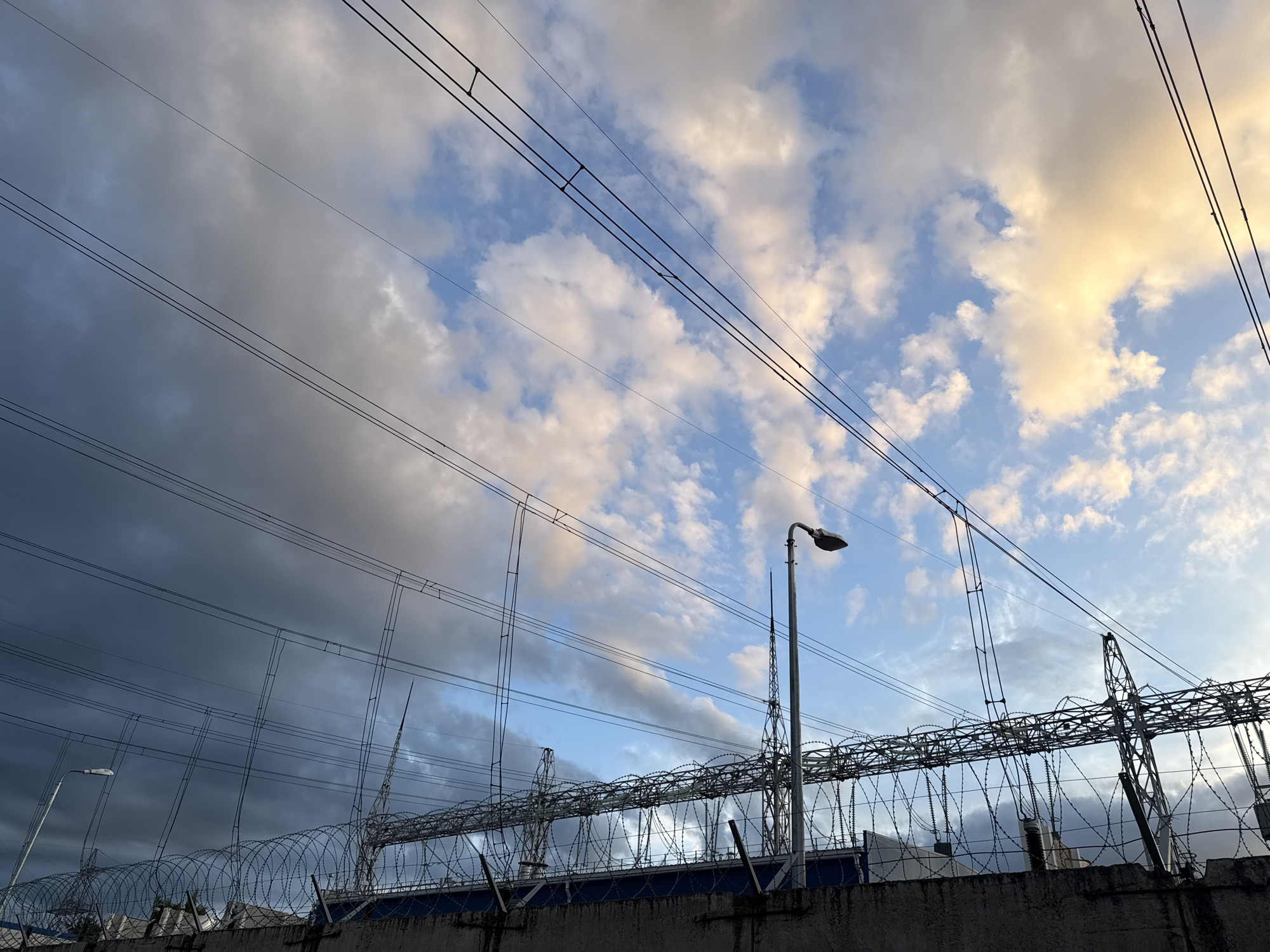 The image shows a high-voltage electrical substation in Chaikovsky, Russia. Visible are power lines stretching across the sky, connected to metal structures and insulators. Below, a concrete wall topped with barbed wire surrounds the facility. A streetlight stands adjacent to the substation. The sky is filled with clouds, with some areas appearing bright blue and others dark gray.