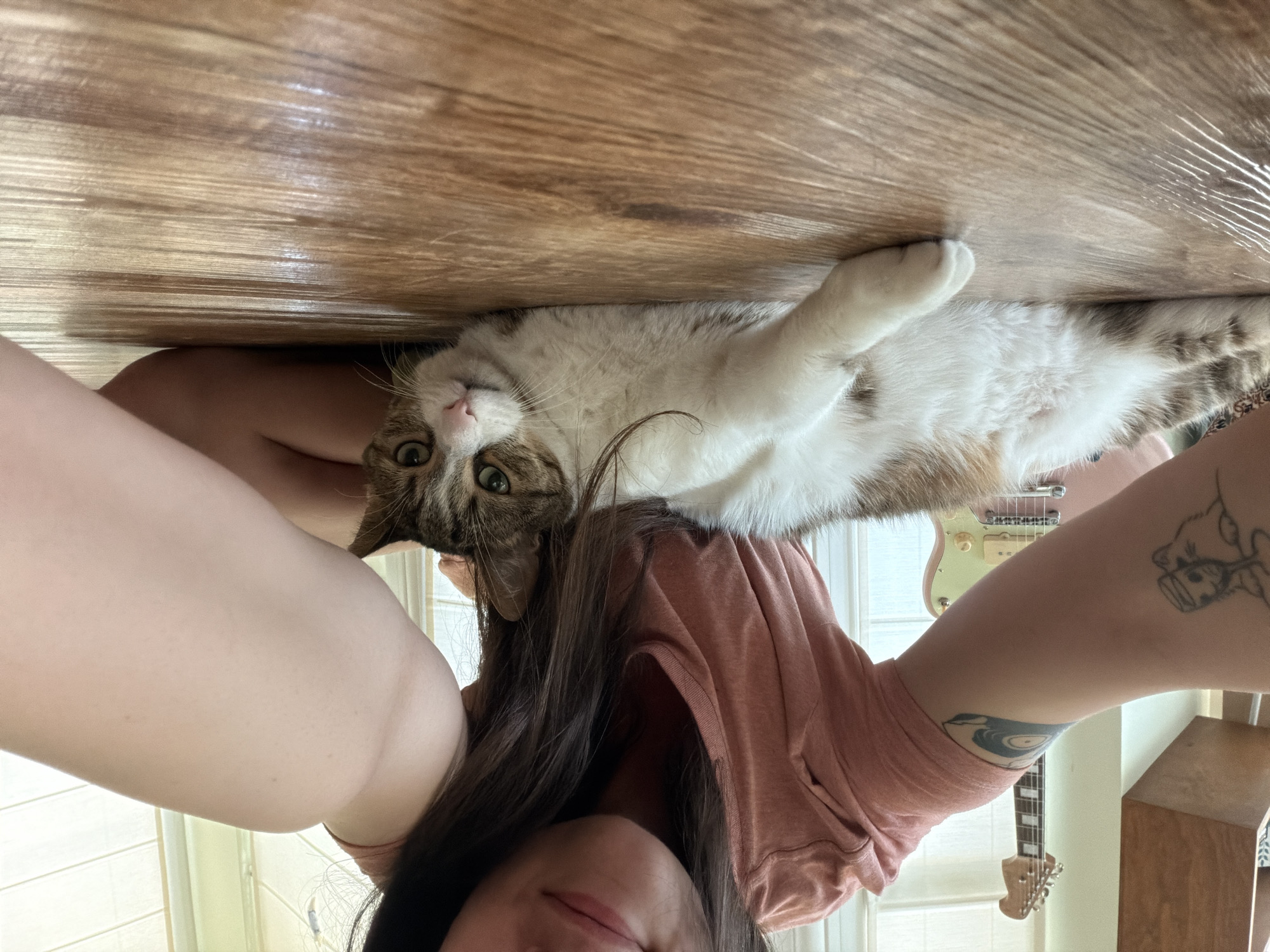 In an indoor setting, likely a residence, a young woman is taking a selfie with her cat. The woman is wearing a pink t-shirt and has long, dark hair. She is positioned on the floor, with her arm extended to capture the photo. To her right, a tabby and white cat is lying down, looking towards the camera. The cat has green eyes and a pink nose. In the background, a guitar is visible, leaning against a wall. The floor appears to be made of wood. The scene suggests a casual and relaxed moment between the woman and her pet.
