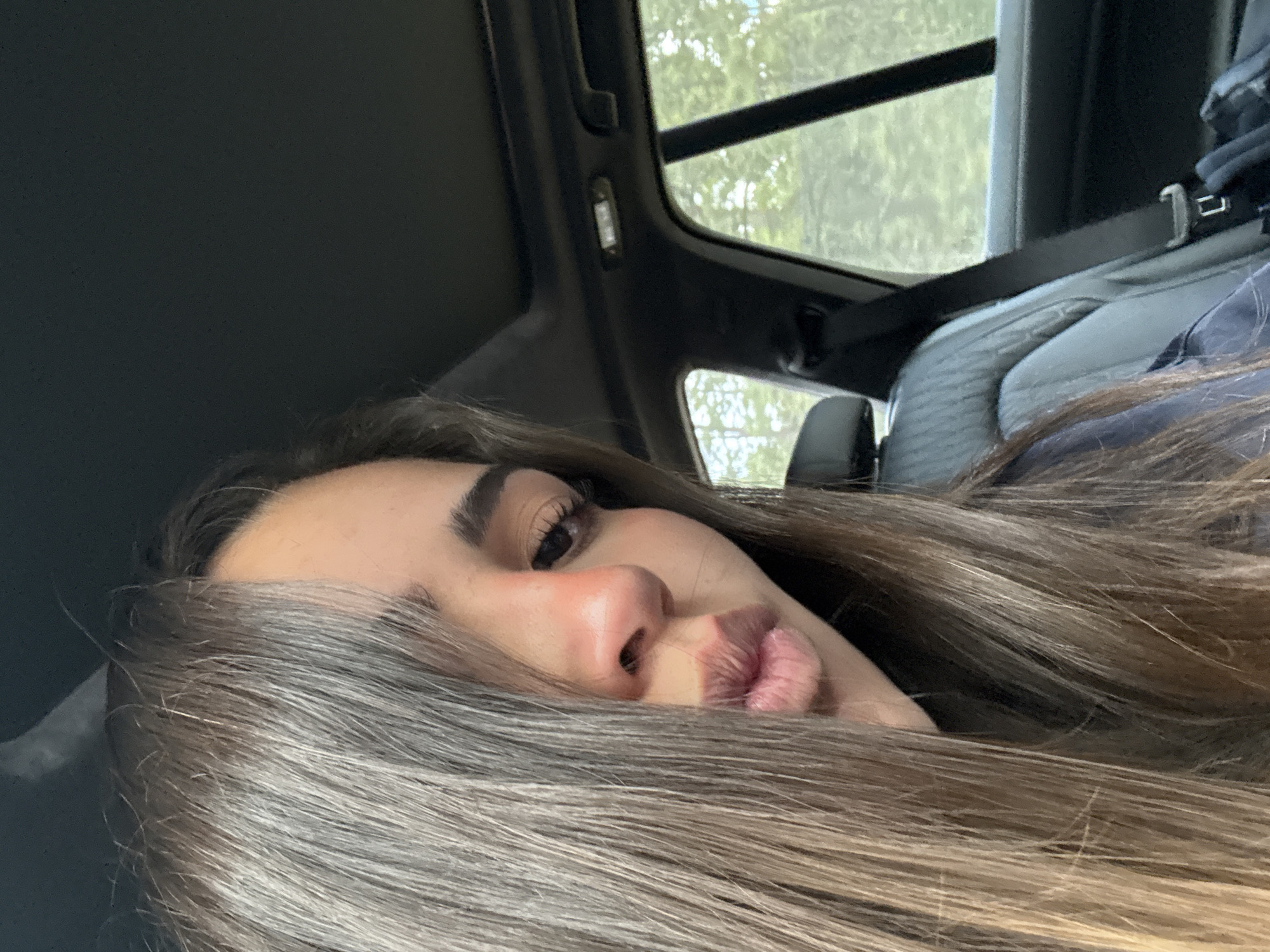 The image shows a woman with long, light brown hair lying in a car. She has brown eyes, full lips, and a light complexion. The car has a sunroof, and trees are visible through the window. The interior of the car is mostly dark gray and black. The seatbelt is visible. The location is not definitively determinable as Rancho Viejo, Mexico, without further contextual clues within the image.