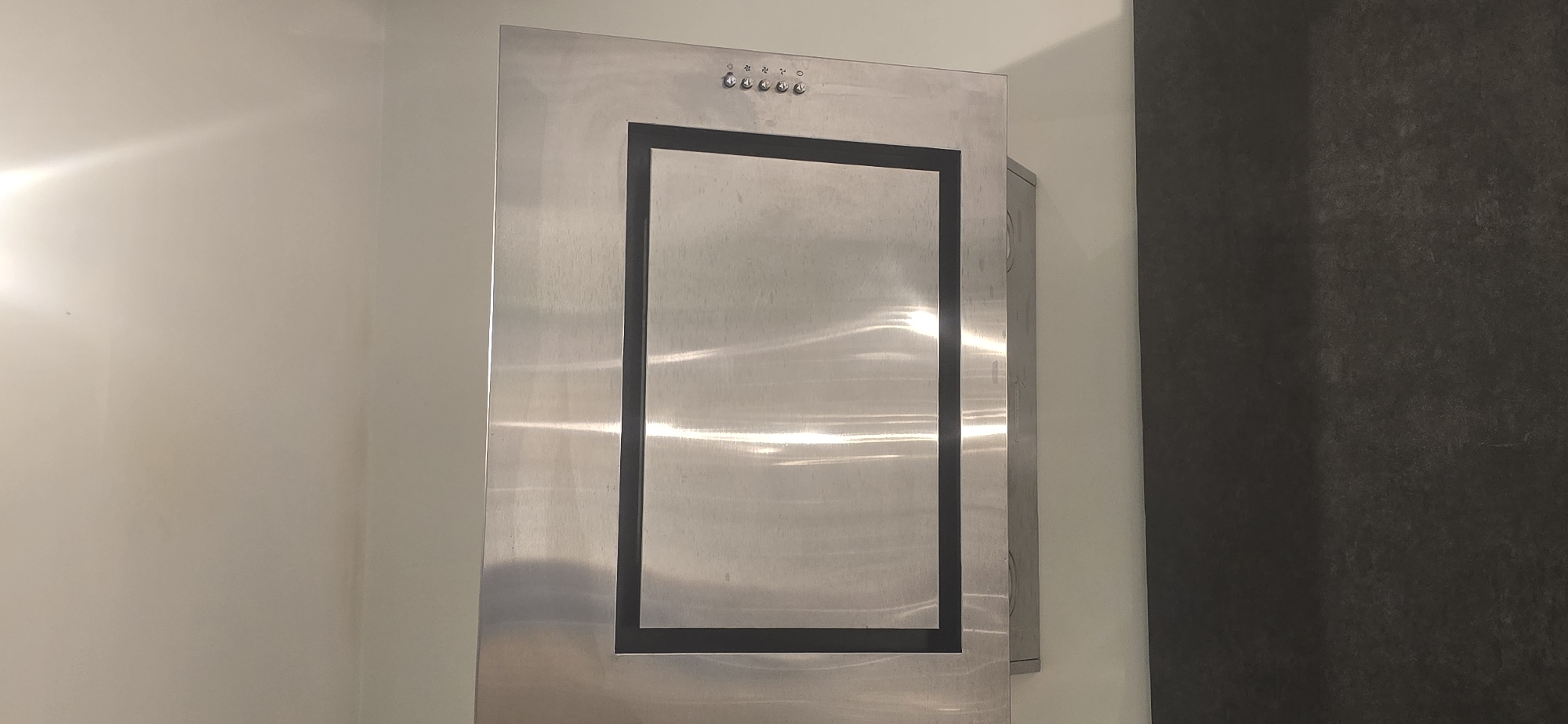 A stainless steel range hood with a black border is mounted on a light-colored wall. The range hood has a rectangular glass panel in the center, and a set of control buttons on the right side. Below the range hood is a dark countertop. The image appears to be taken in a kitchen. The location is Unknown, Unknown.