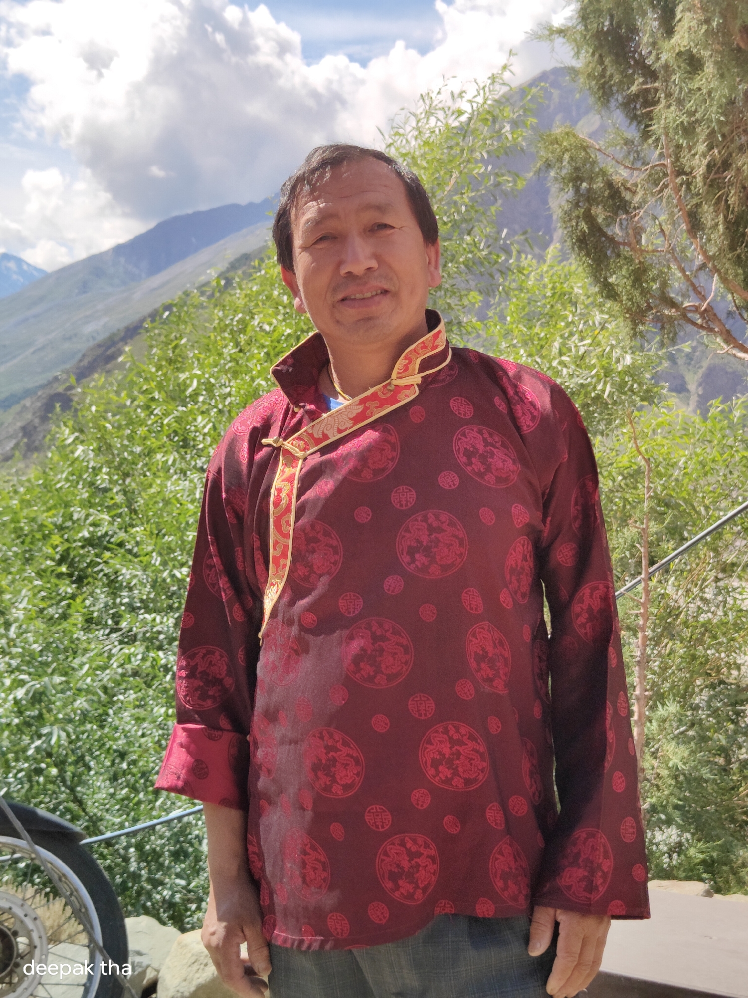 An Indian man, appearing to be in his late 50s, stands outdoors, facing the camera. He has short, dark hair, a medium complexion, and is smiling. He is wearing a traditional maroon silk tunic with a Chinese dragon pattern in a darker shade of red, accented with gold trim around the collar and down the front opening. The tunic has a high collar and is fastened with a knot at the neck. He is also wearing a blue shirt underneath and blue jeans.
He is standing in front of a backdrop of lush green foliage, with mountainous terrain and a cloudy sky visible in the distance. The setting suggests a rural or mountainous area, consistent with locations like Darcha, India, which is known for its mountainous landscape. In the lower left corner of the image, a portion of a bicycle wheel is visible, suggesting it may be nearby. The overall lighting indicates it is daytime, likely sunny given the bright sky and shadows. The image appears to be a candid portrait.