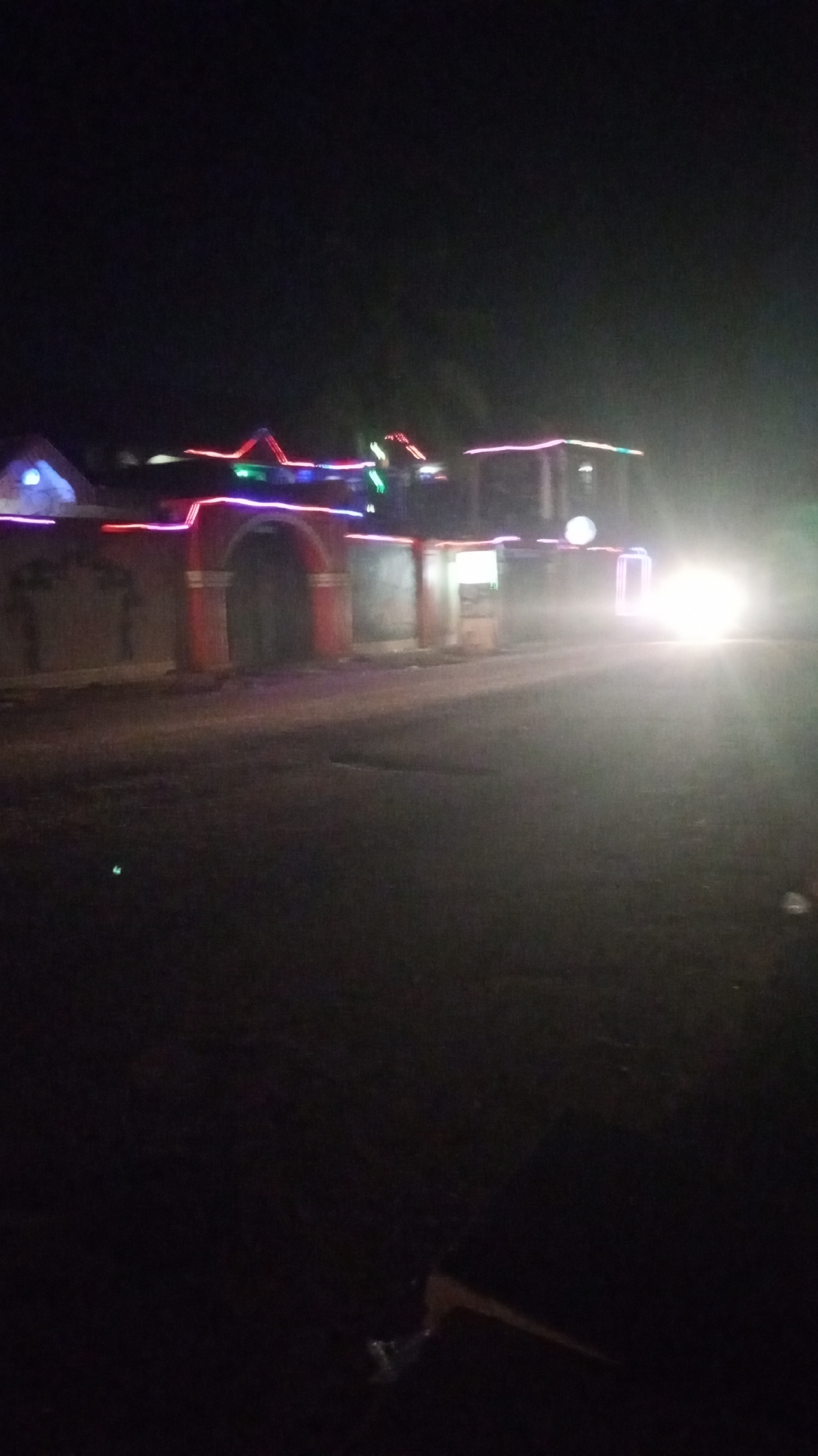At night in Sabon Tasha, Nigeria, a brightly lit vehicle approaches on a road. Buildings are decorated with colorful lights. The architecture is a mix of residential and commercial, featuring arched entrances and walled perimeters. The road surface appears paved, with visible imperfections. The sky is dark, suggesting limited ambient light.
