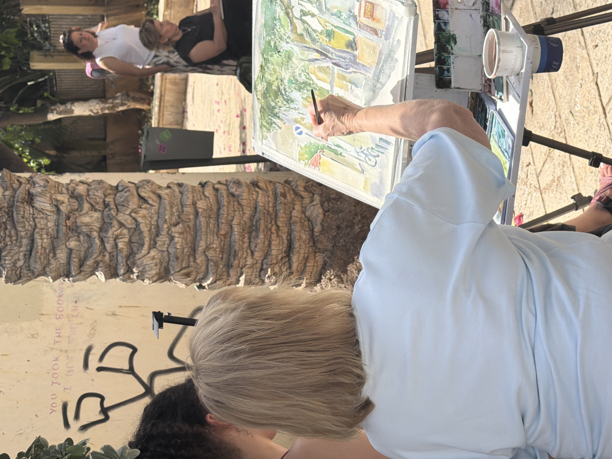 This image captures an outdoor art scene on a sunny day in Tel-Aviv, Israel. The primary subject is a person with blonde hair, seen from behind, wearing a light blue short-sleeved shirt. They are actively painting on an easel with a brush, creating a watercolor landscape that depicts a street scene with buildings, trees, and a blue circular road sign.
Next to the painting, on the easel's stand, a palette of watercolors is visible along with a white cup, partially showing the text "toshida" and a blue stripe. The ground beneath is paved with light-colored stones or tiles.
In the mid-ground, two other individuals are present. One person, wearing a white t-shirt and dark sunglasses pushed onto their head, stands or sits further back. Another person, dressed in a black shirt, appears to be relaxing or observing behind them. On the left side of the frame, a prominent, textured palm tree trunk suggests the warm, Mediterranean climate.
Behind the painter, a light-colored wall is covered with graffiti. Visible text reads "YOU TOOK THE BOOK" and "I GOT THE CH-", accompanied by abstract black spray-painted shapes. A black speaker with a small green and pink logo is also present near the wall. The bright sunlight indicates daytime, casting strong shadows and highlighting the vibrant outdoor activity.