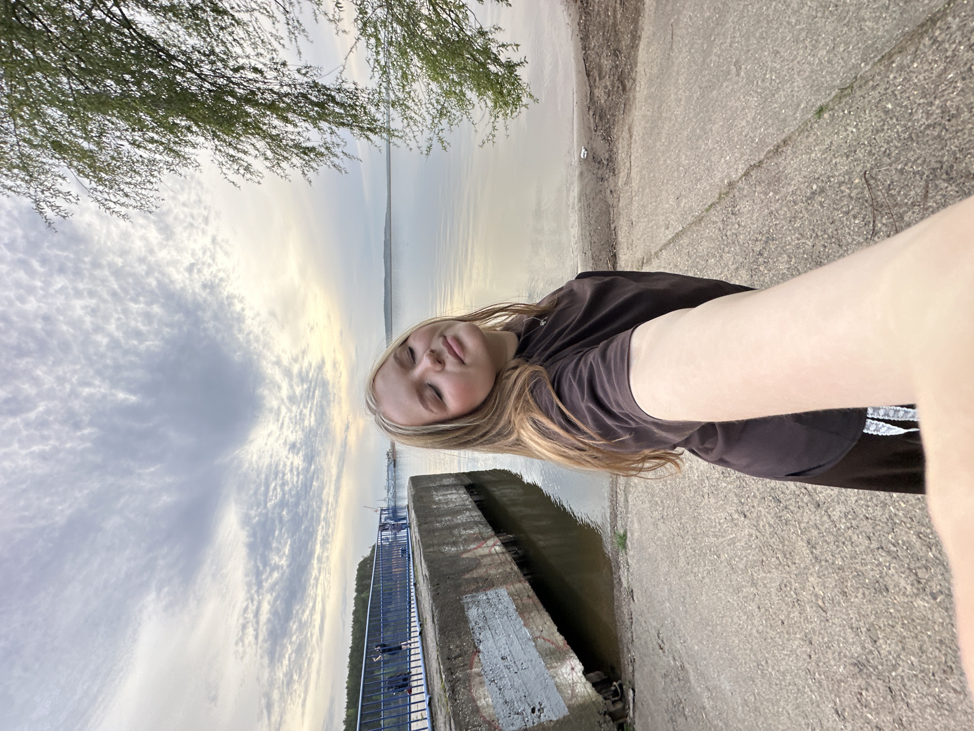 A selfie-style photograph shows a young woman with light blonde hair, wearing a dark t-shirt and shorts, standing outdoors in the city of Chaikovsky, Russia. The photograph is rotated 90 degrees counterclockwise. The woman's face is visible, and she appears to be holding the camera at arm's length. In the background is the Kama River, with a cloudy sky above. A concrete embankment runs along the river's edge. A blue railing can be seen on a section of the embankment, and a concrete structure extends into the water. In the upper left corner of the image is a tree branch with green leaves.
