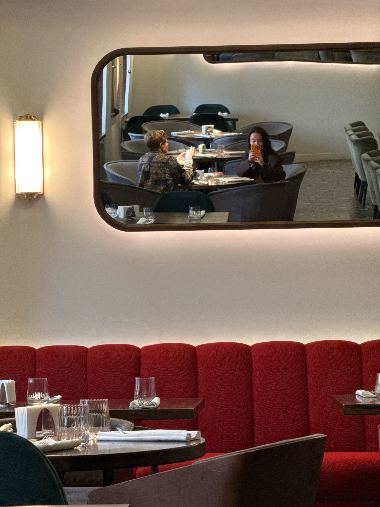 The image depicts the interior of a restaurant with a modern, slightly upscale ambiance. A large, horizontally oriented mirror with rounded corners is mounted on a cream-colored wall. The mirror reflects a portion of the restaurant's seating area. In the reflection, two women are seated at separate tables. The woman on the left is wearing a patterned shirt and is looking down at her phone. The woman on the right is wearing a dark jacket and is also looking at her phone, which is bright orange. Both women appear to be dining alone. The mirrored area reveals several dark-colored, plush chairs arranged around circular tables, some of which are set with glassware and napkins. A wall-mounted lamp with a fluted glass shade and brass accents is visible on the left side of the actual image, casting a warm light. Below the mirror and the wall, a section of curved red booth seating is prominent, with several small, dark wooden tables in front of it. These tables are also set with glasses and napkins, suggesting a dining setting. The overall impression is a well-lit, contemporary dining space, potentially a restaurant in Novaya Usman', Russia, given the context.