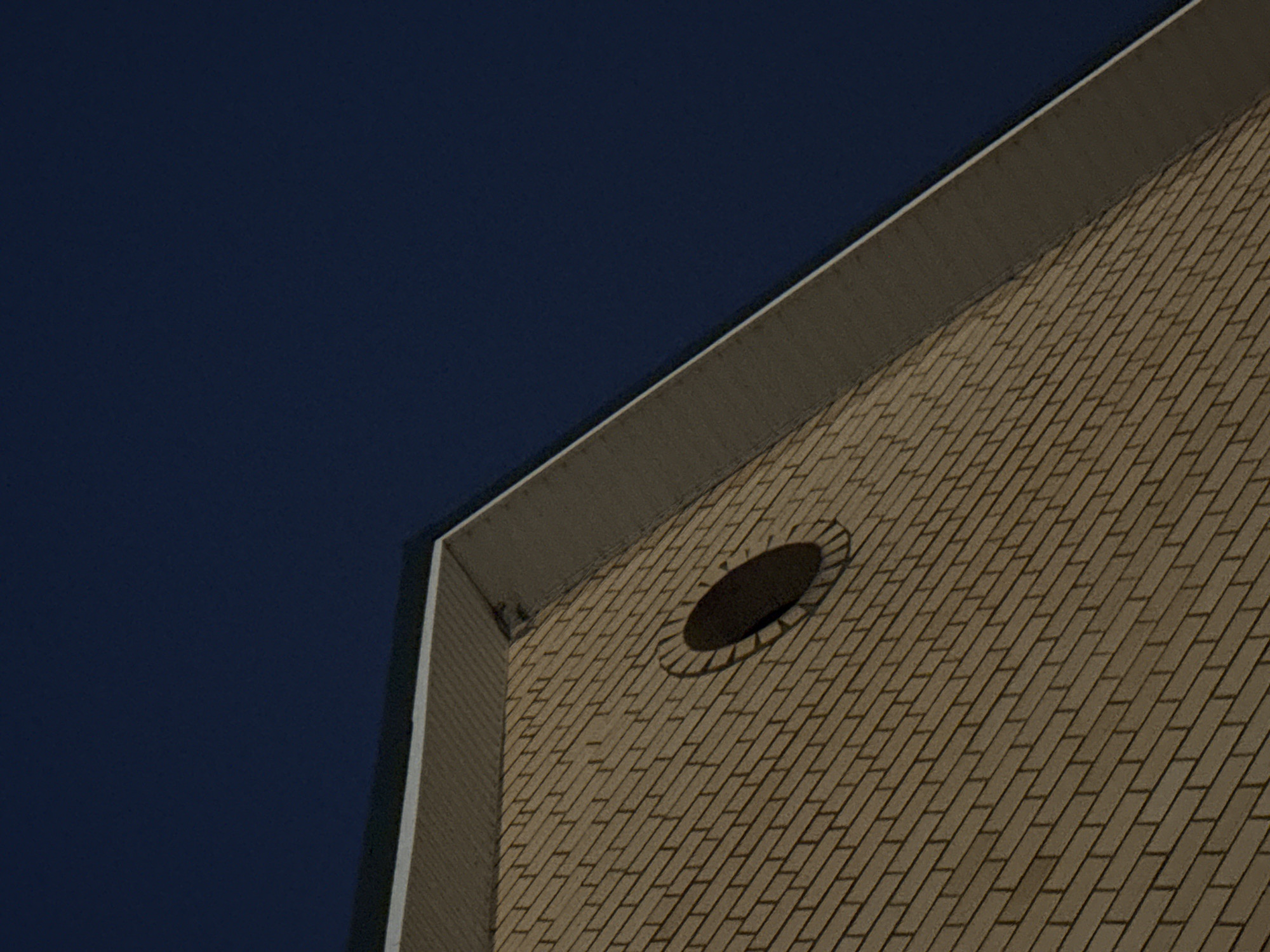 This is a low-angle, medium-shot photograph of the side of a brick building against a dark blue sky. The building's facade is constructed of light brown bricks laid in a stretcher bond pattern. A white soffit lines the eaves of the roof, which is angled towards the upper right of the frame. Centrally located on the brick wall is a dark, circular window with a decorative brick surround. The image is part of a larger dataset of photographs taken in the city of Zaokskiy, Russia.