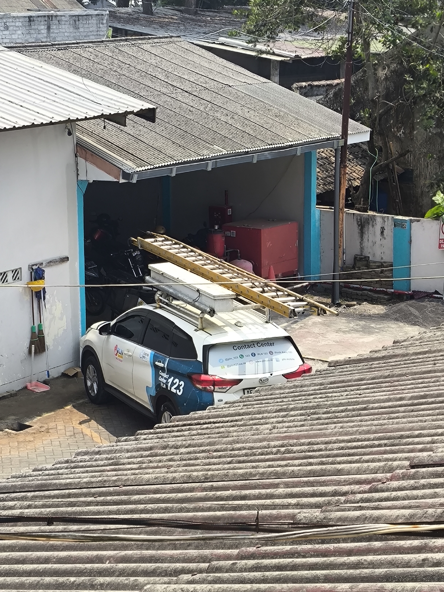In Sukodono, Indonesia, a white SUV with "Contact Center PLN 123" branding is parked outside a building with a corrugated metal roof. The vehicle is equipped with a roof rack carrying a ladder and other equipment. To the left of the vehicle, cleaning supplies including brooms and a bucket are leaning against the building's white wall. The building appears to be a garage or workshop, as motorcycles are visible inside. The scene is outdoors with visible overhead power lines and trees in the background. The ground is paved with interlocking bricks.